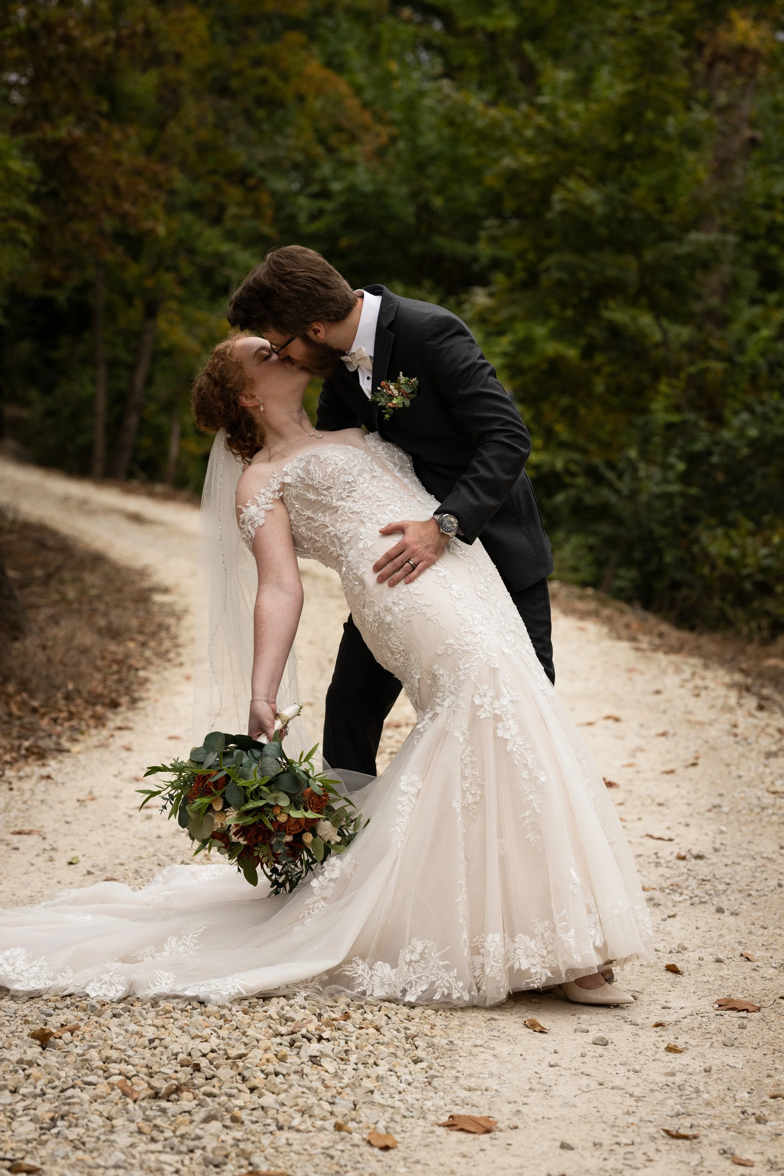 A bride and groom share a kiss on a dirt path surrounded by trees during a wedding photoshoot, with the groom dipping the bride and holding her waist, she holding a bouquet of flowers.