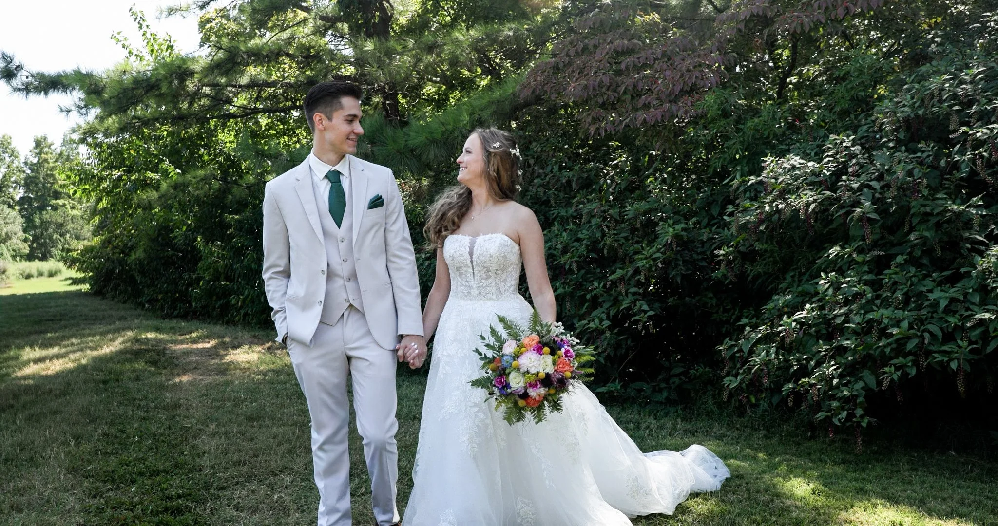 A bride and groom walk hand in hand outdoors, surrounded by greenery. The bride is wearing a white wedding gown and holding a colorful bouquet, while the groom is dressed in a light-colored suit with a green tie. They are smiling at each other in a garden setting.