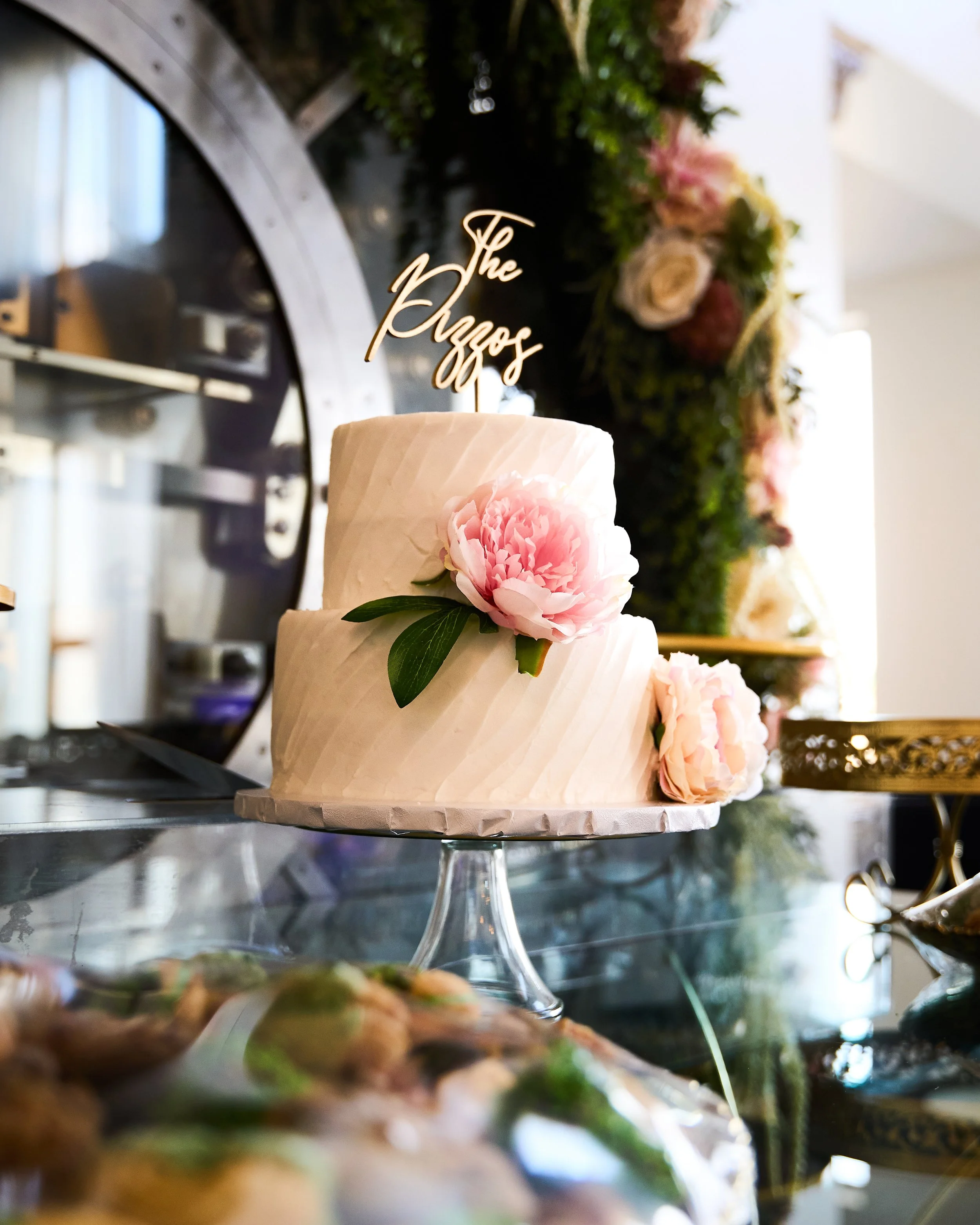 A tiered cake with pink flowers and green leaves, topped with a 'The Pozzos' cake topper, placed on a glass cake stand at a celebration or event.