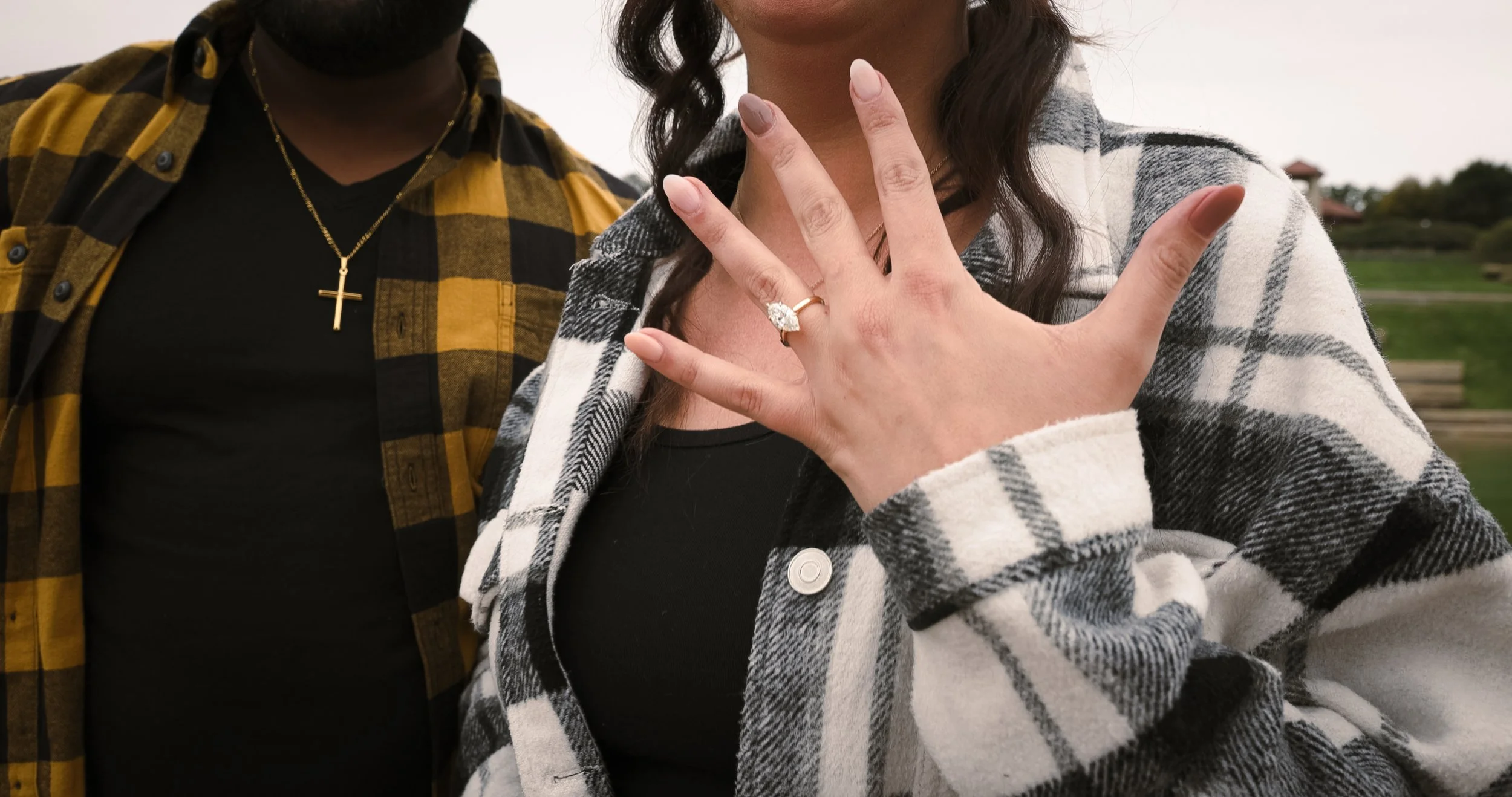A woman showing off her engagement ring on her left hand, with a man standing beside her outdoors.