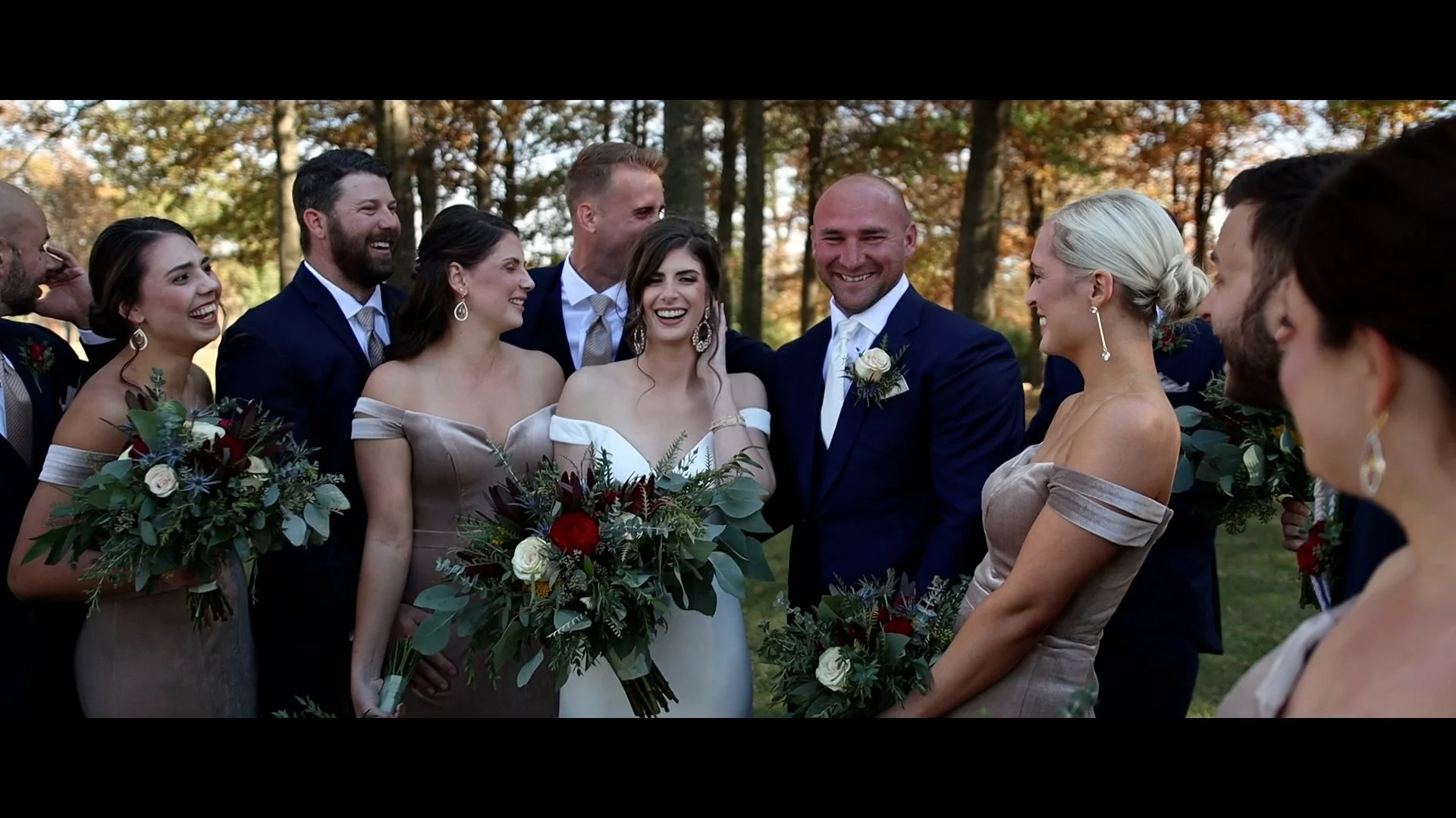 A wedding party outdoors with the bride, groom, bridesmaids, and groomsmen smiling and holding bouquets, surrounded by trees with autumn foliage.