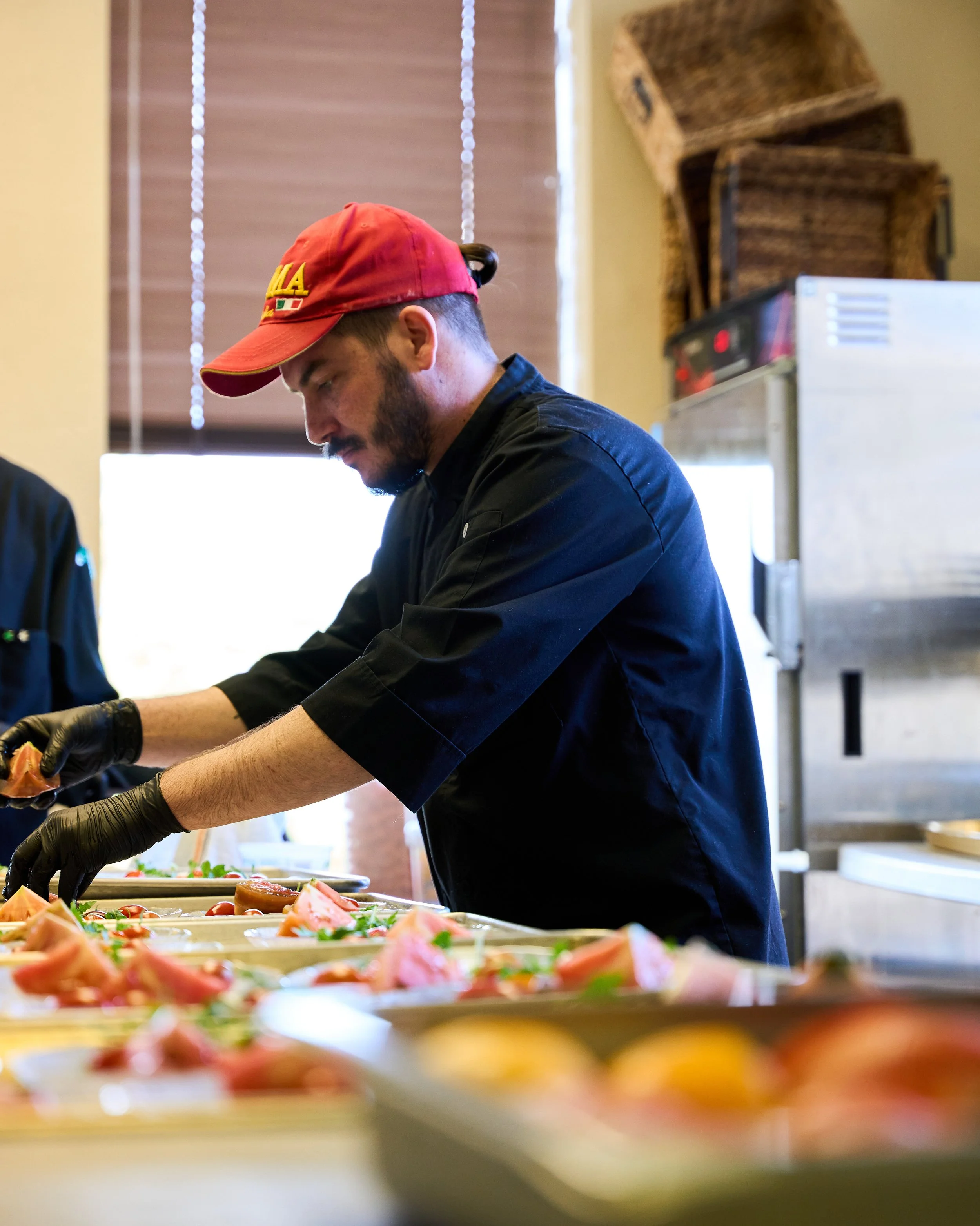 A male chef with a beard wearing a black chef jacket, black gloves, and a red baseball cap, preparing food in a kitchen.