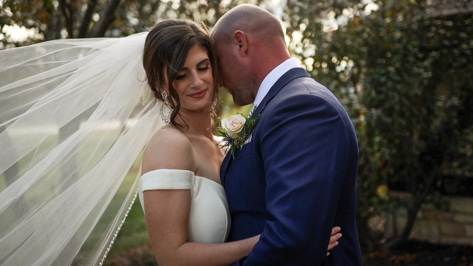 Bride and groom embracing outdoors during wedding, bride wearing off-the-shoulder white dress and veil, groom in navy suit with boutonniere, tree background.