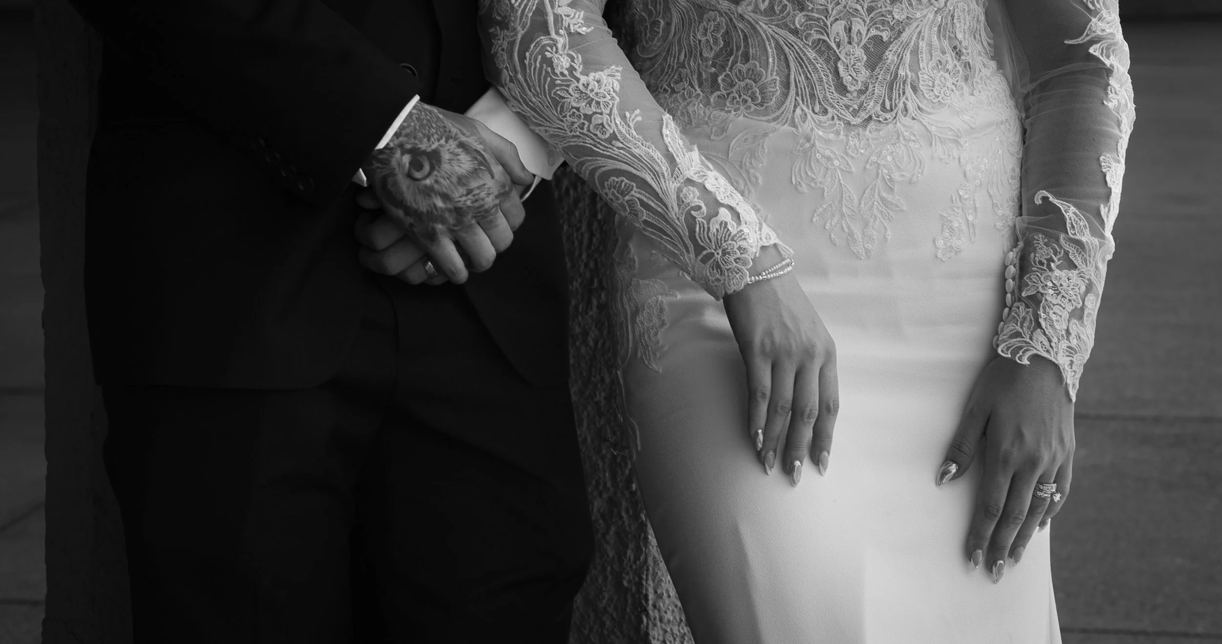 A bride and groom hold hands, with the bride wearing an ornate lace wedding dress and the groom wearing a dark suit. Their hands are intertwined, and the bride's ring and manicure are visible. The photo is in black and white.