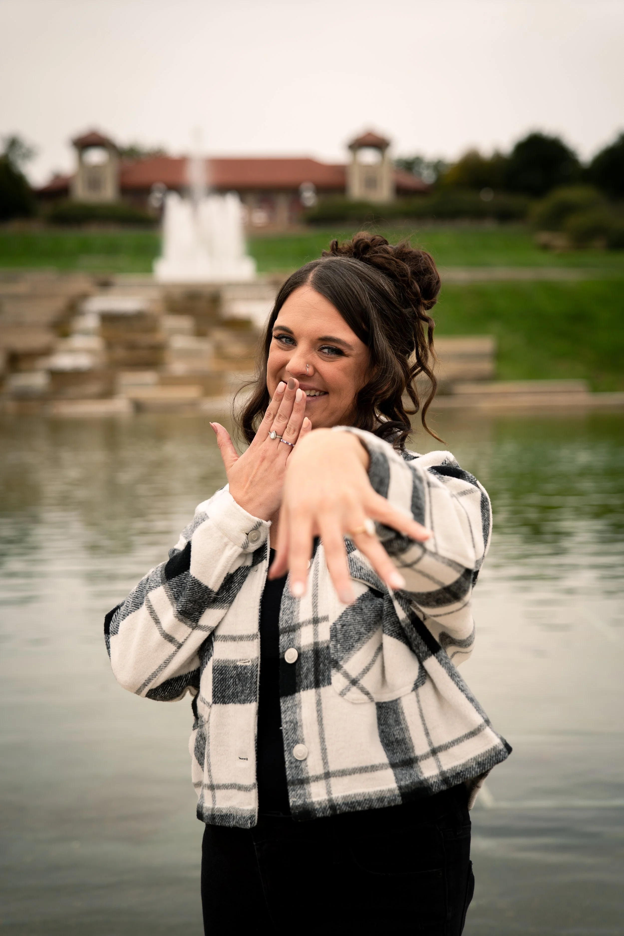 Smiling woman with dark curly hair in a plaid jacket making a playful gesture near water with a park and fountain in the background.