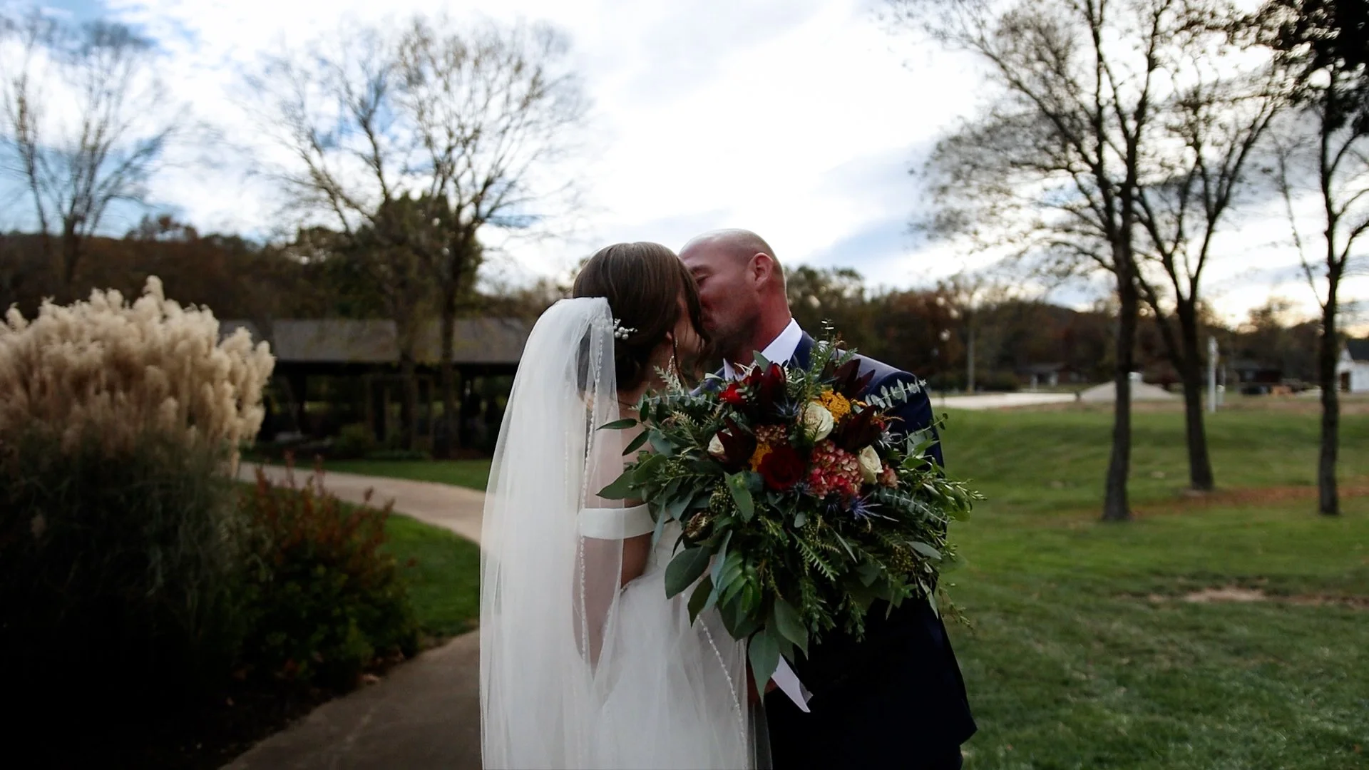 A bride and groom sharing a kiss outdoors on a cloudy day, with the bride holding a large bouquet of flowers, trees, and a pathway in the background.