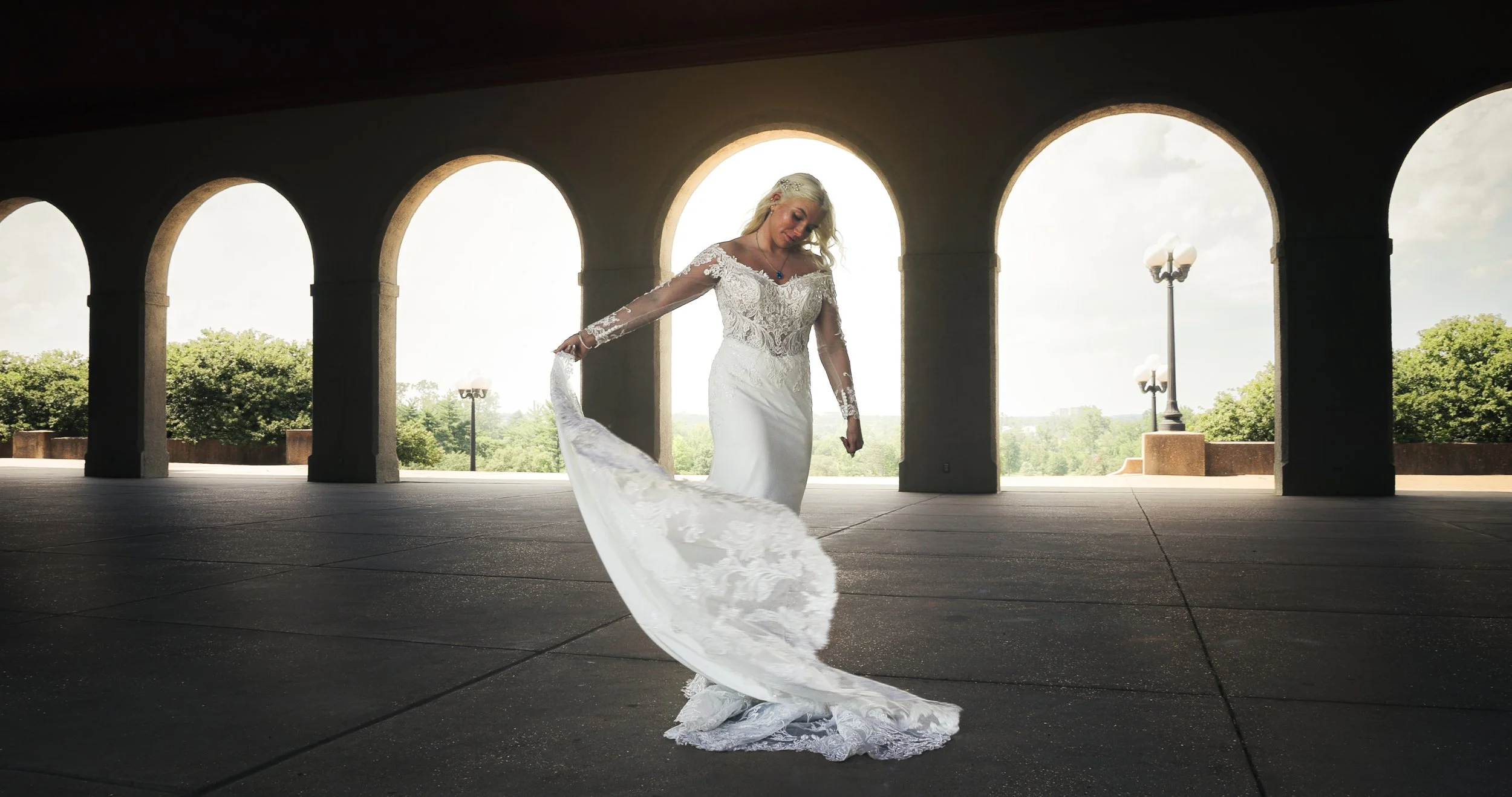 A bride in a wedding dress with lace details, standing under an arched structure with a landscape and trees in the background.