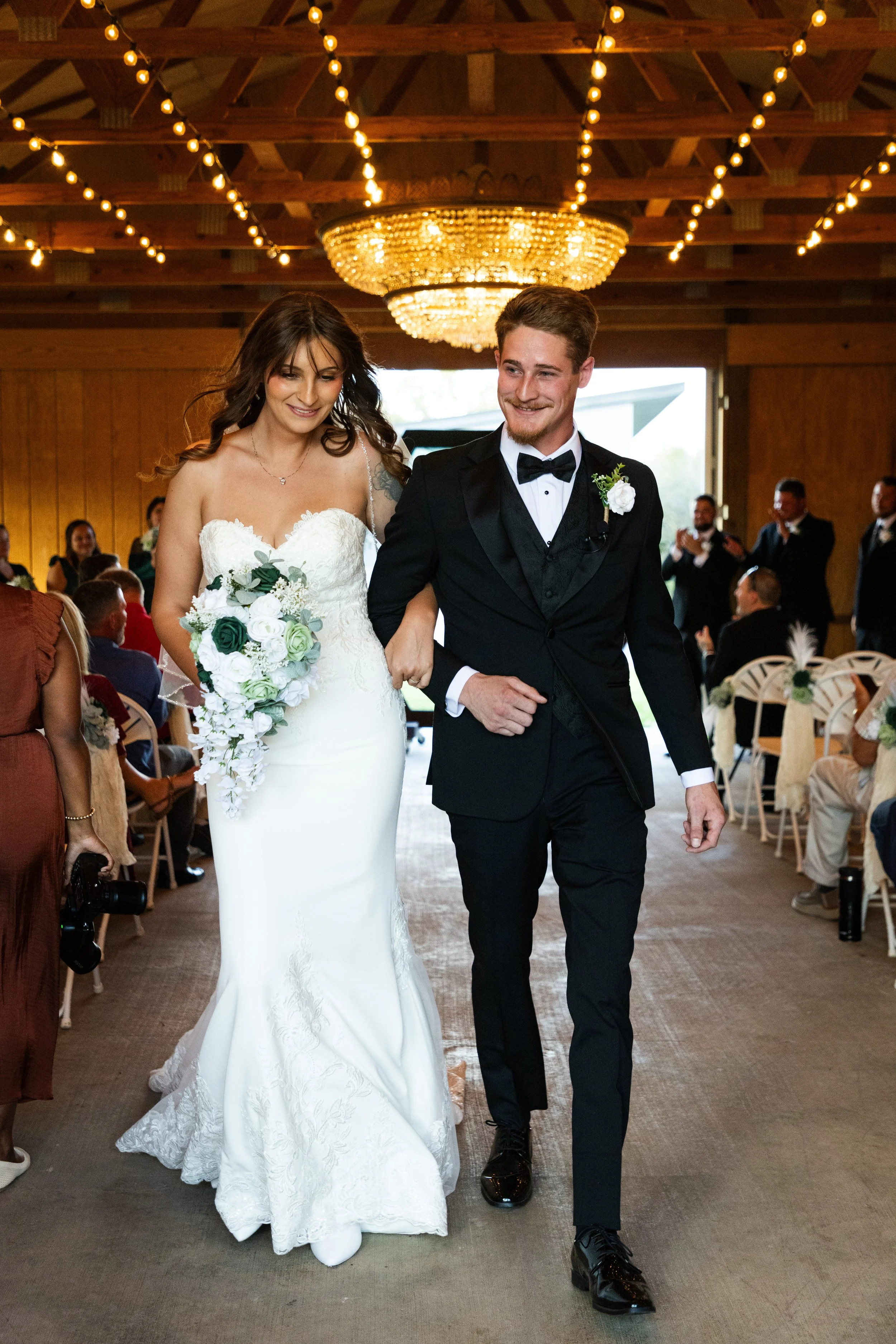 Bride and groom walking down the aisle at a wedding ceremony. The bride is in a white wedding dress holding a bouquet, and the groom is in a black tuxedo. Guests are seated on either side, and there is a chandelier and string lights hanging from the wooden ceiling.