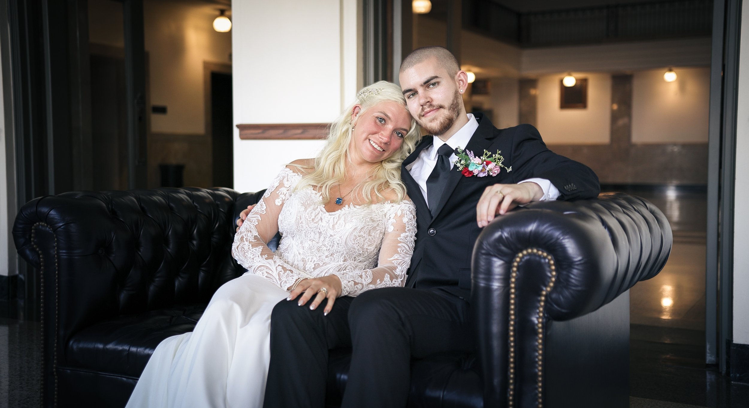 A bride and groom sitting on a black leather sofa, smiling at the camera. The bride wears a white lace wedding dress, and the groom wears a black suit with a white shirt and tie. They are in an indoor setting with warm lighting.