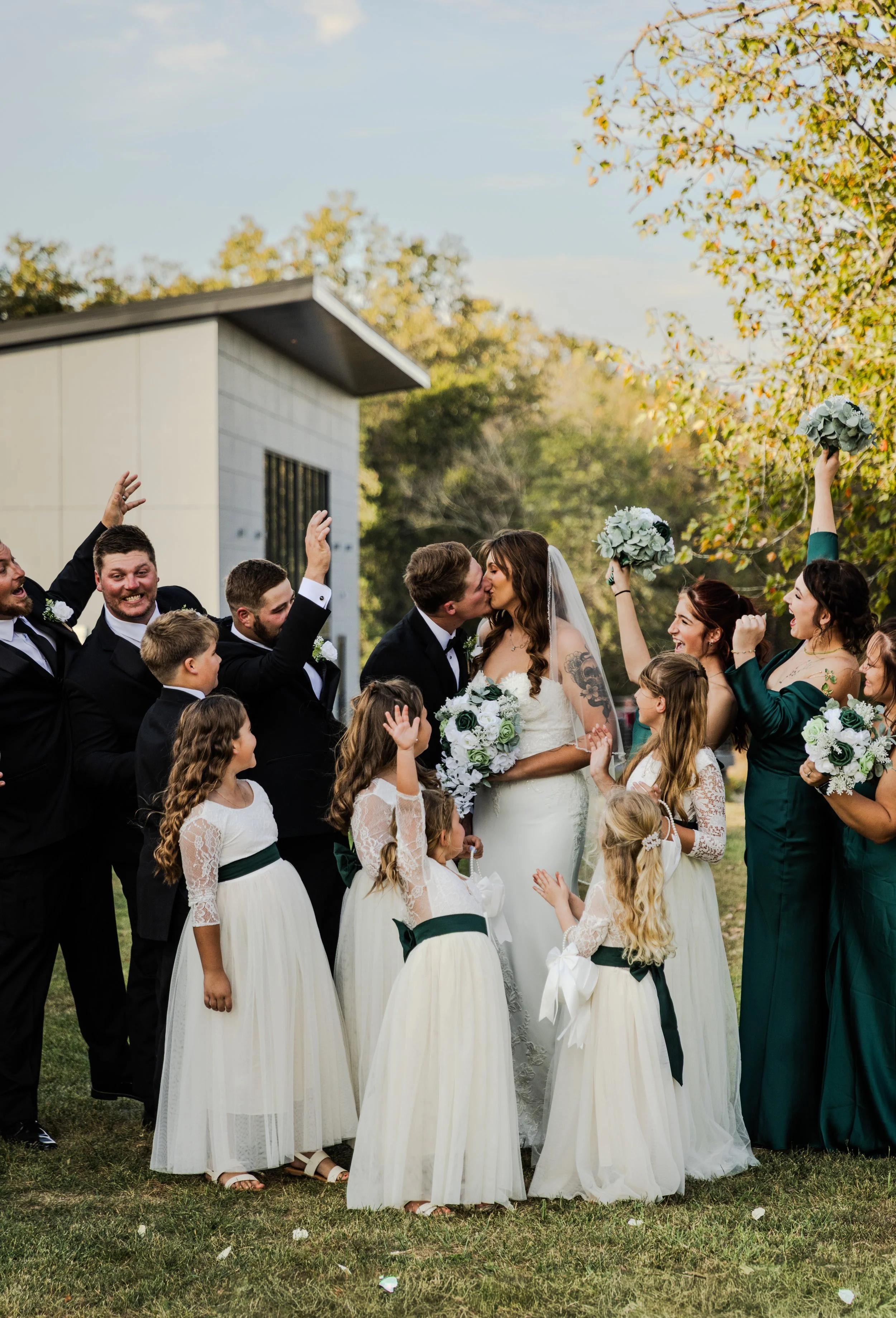 A wedding celebration with a bride and groom kissing, surrounded by bridesmaids, groomsmen, and young flower girls, outdoors on a grassy area with trees and a small building in the background.