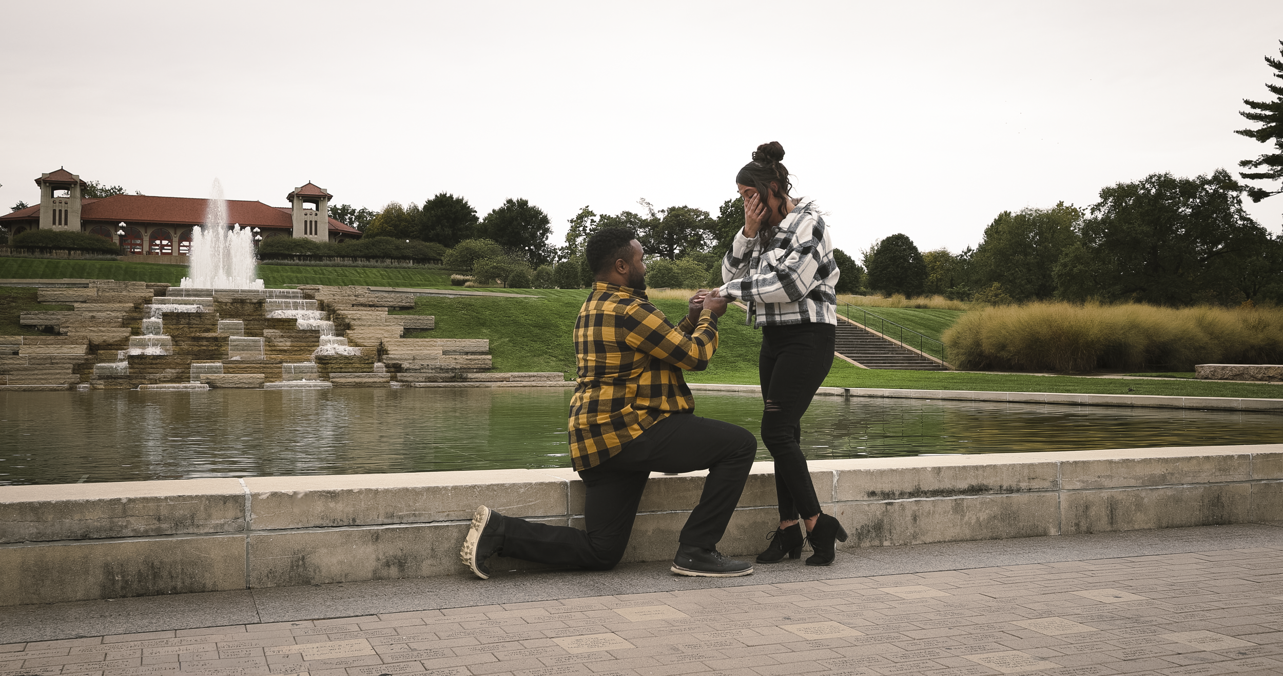 A man is proposing to a woman by kneeling and holding her hand, while she covers her mouth with surprise, in front of a fountain and park scenery.