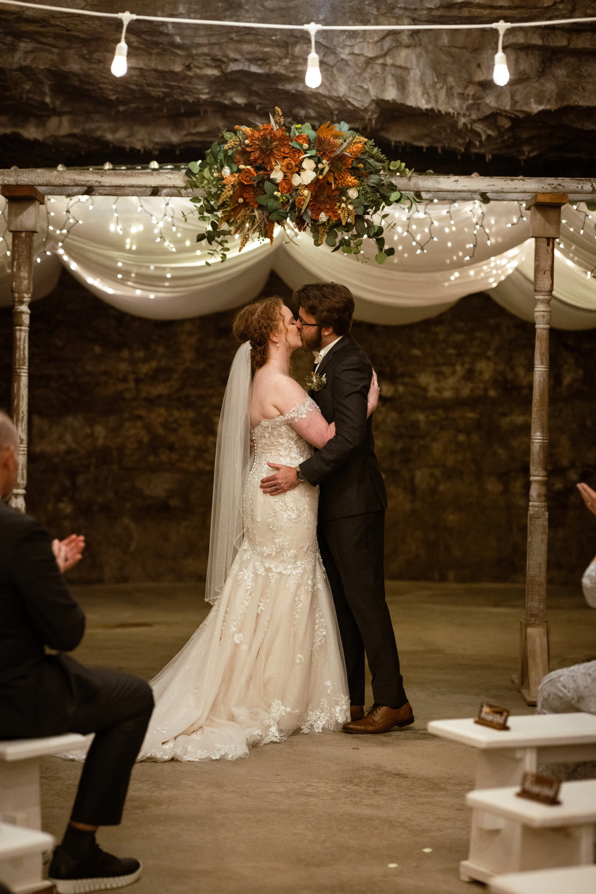 A bride and groom share a kiss on their wedding day, surrounded by guests clapping, under a decorated arch with string lights and a floral arrangement.