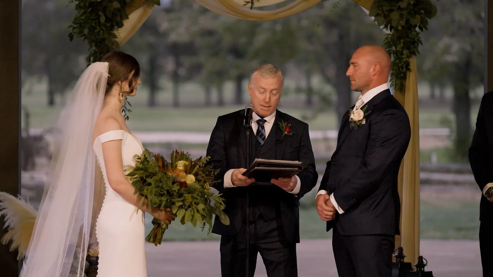 A bride and groom standing at the altar during their wedding ceremony, with an officiant reading from a book. The bride is holding a bouquet of flowers and wearing a white dress with a veil, while the groom is in a dark suit with a boutonniere. The ceremony is outdoors with a scenic background of trees and water.
