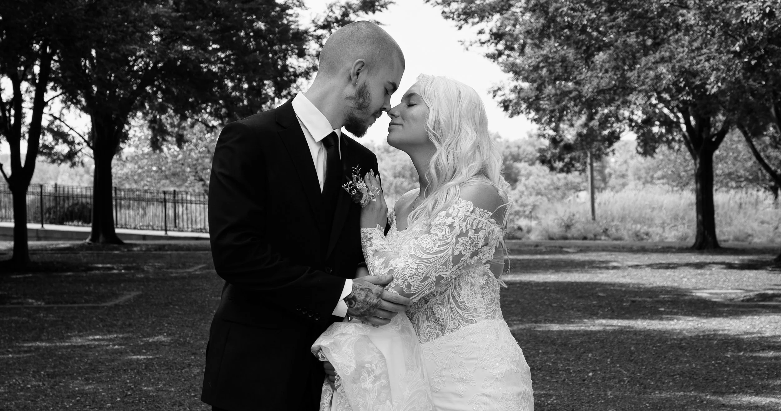 A bride and groom stand close together in an outdoor park, touching their foreheads and gazing into each other's eyes, both with serene expressions. The scene is in black and white, with trees and a fence in the background.