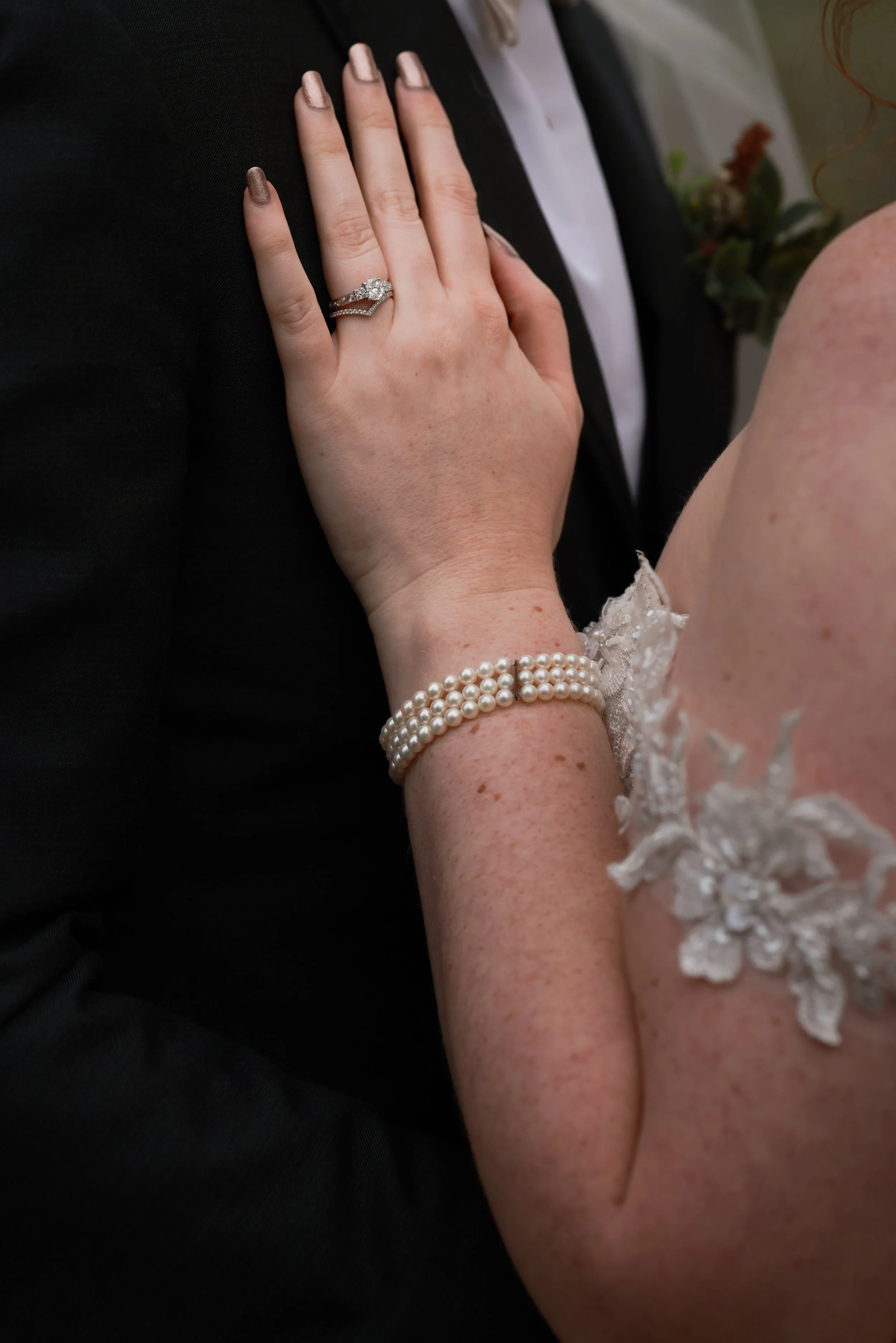 A bride wearing a pearl bracelet and floral lace wedding gown gently touching the chest of a groom in a black tuxedo.