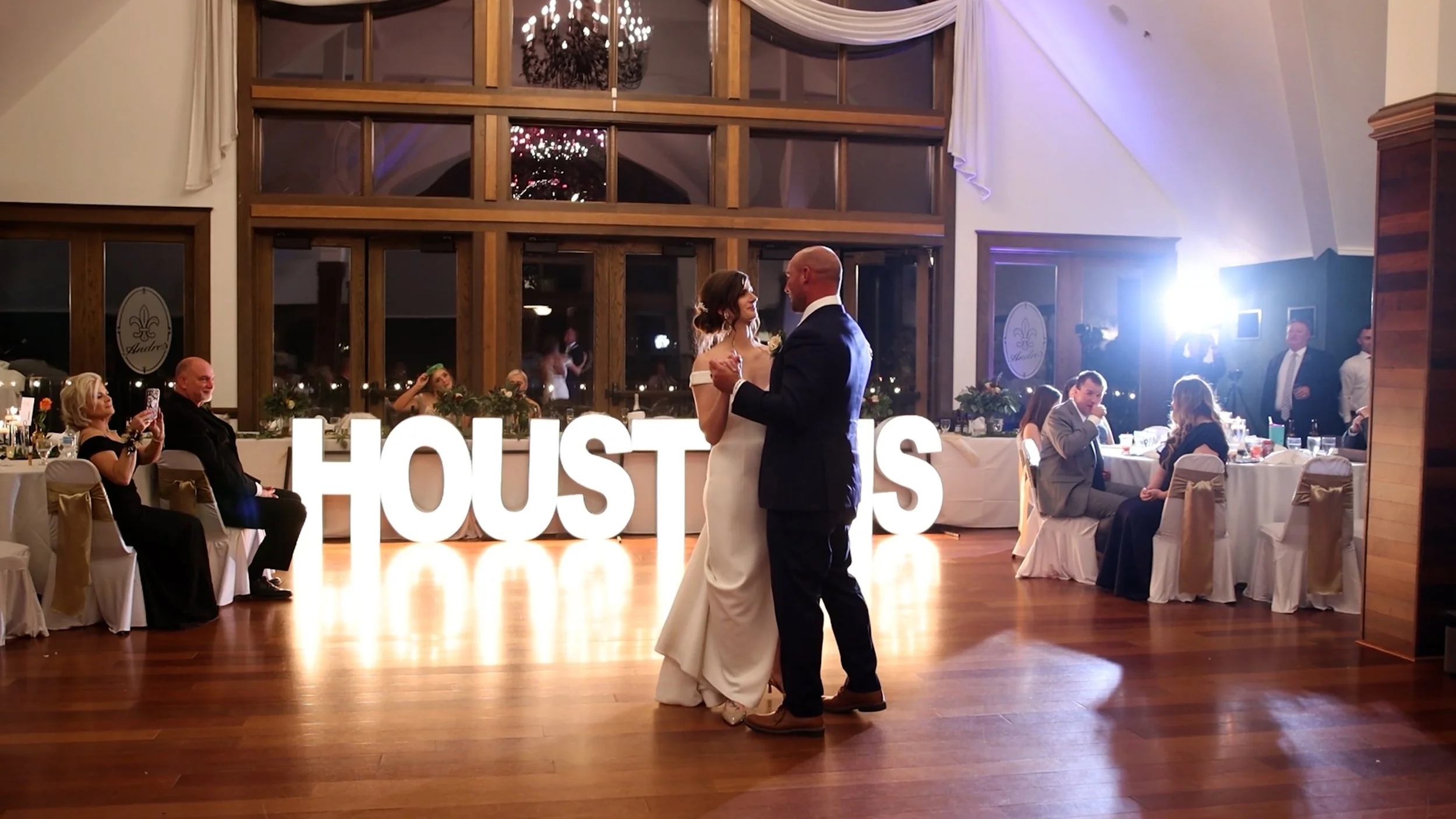 A bride and groom sharing a dance at their wedding reception in a decorated hall with guests seated at tables and taking photos. Large illuminated letters spell out 'HOUSTON'S' in the background.
