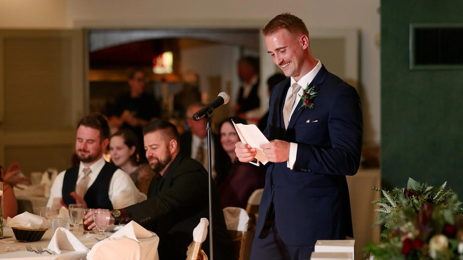 A man in a blue suit giving a speech at a wedding reception, standing at a microphone with friends sitting at a table behind him.