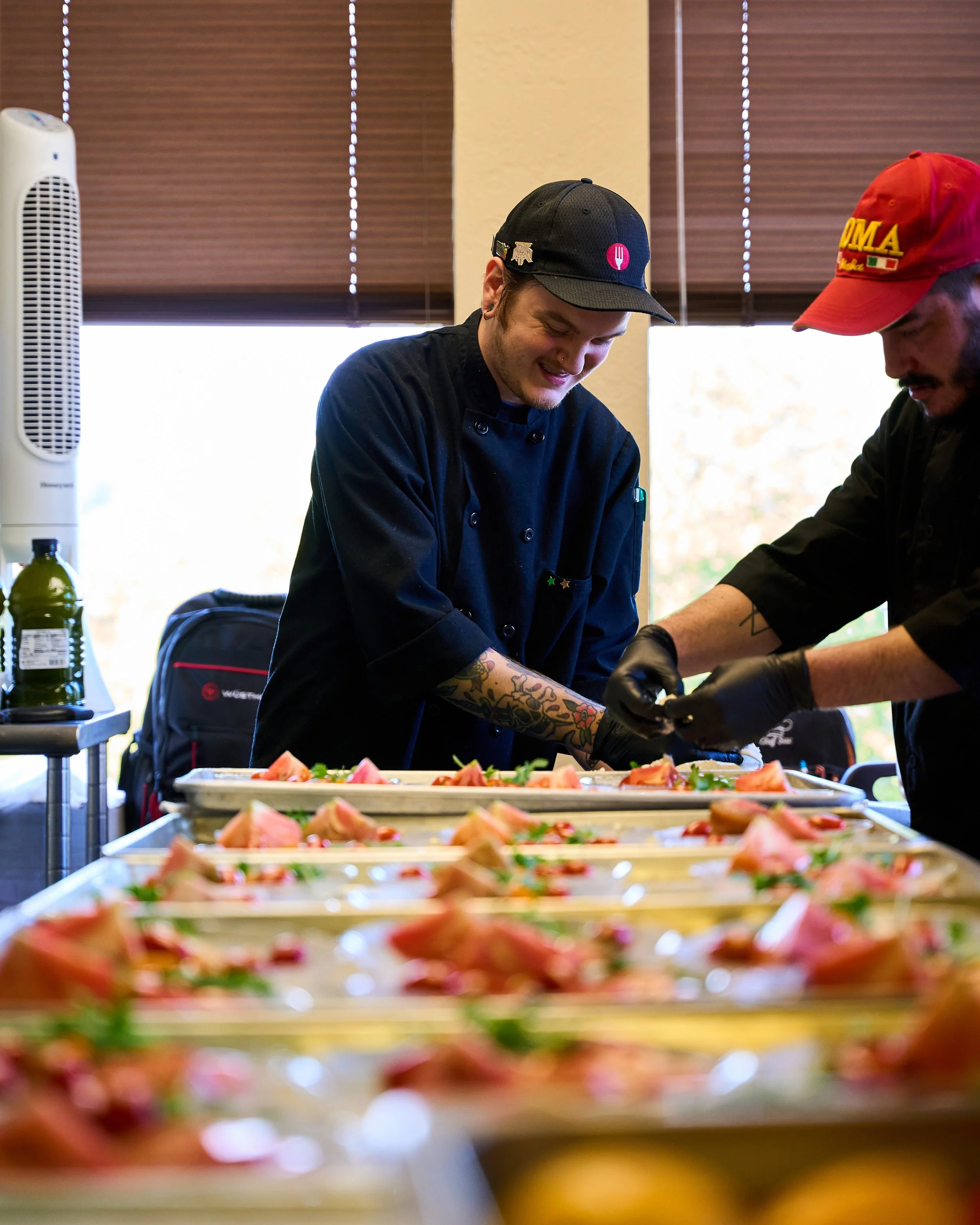 Two chefs preparing plated dishes in a kitchen with trays of food, kitchen knives, and a backpack, window with blinds in the background.
