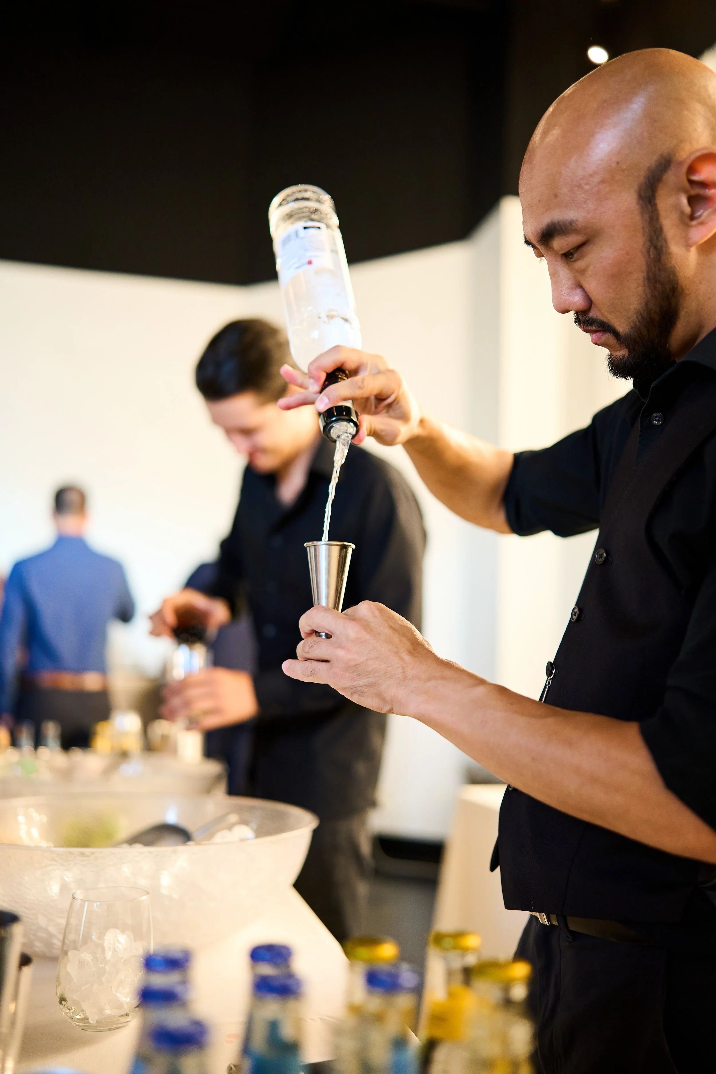 Two bartenders preparing drinks at a bar, with one pouring a clear liquid from a bottle into a shaker and the other working with ingredients in the background.