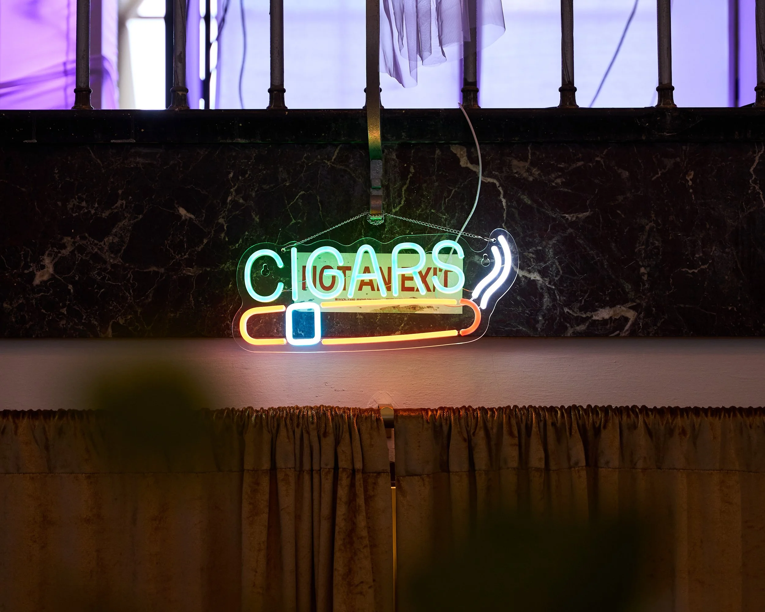 Neon sign saying 'CIGARS' with an image of a smoking cigarette, hanging on a dark marble wall above a curtain.