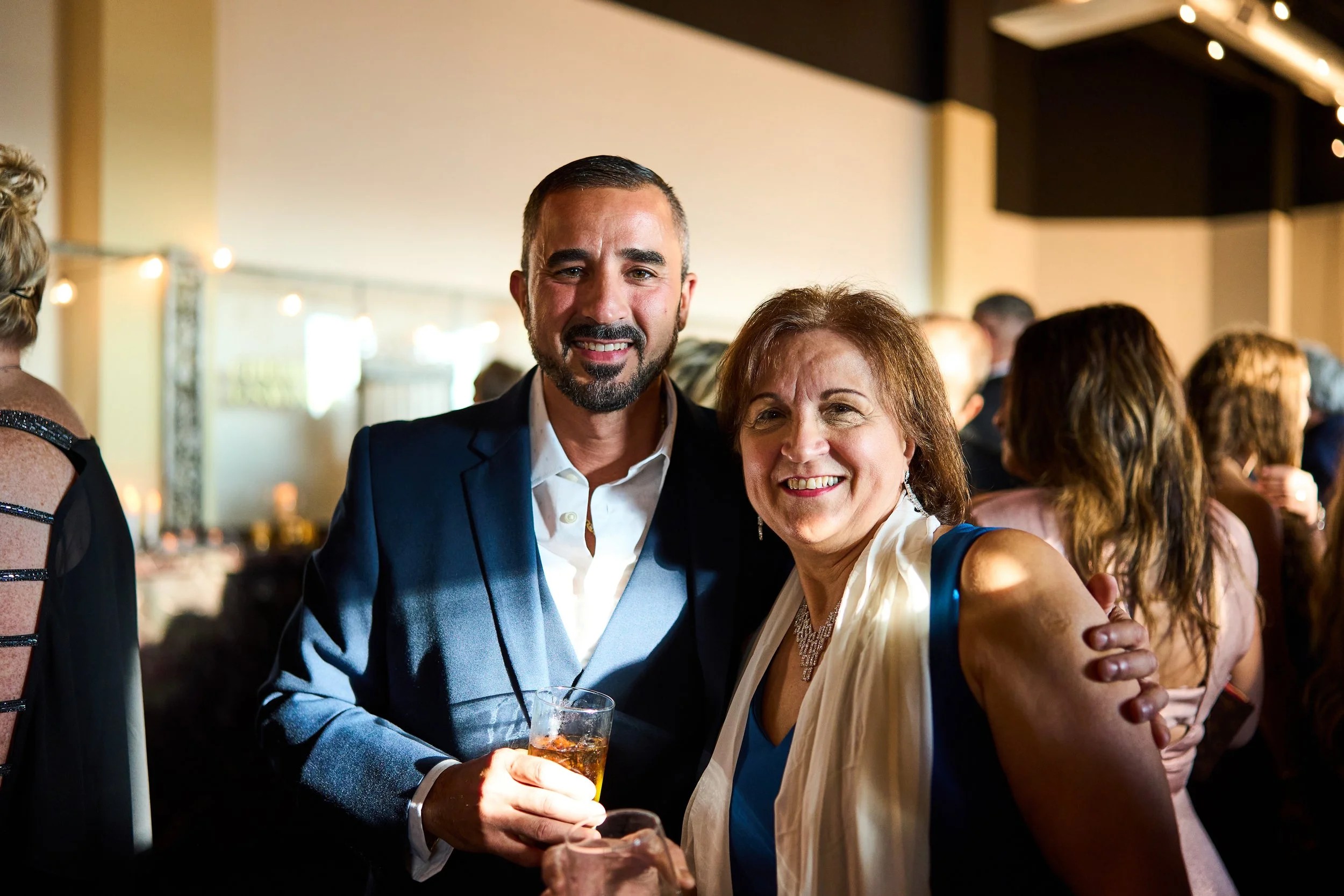 A man and a woman smiling at a social event, with the man holding a drink. They are dressed in formal attire, standing close together, with other guests in the background.