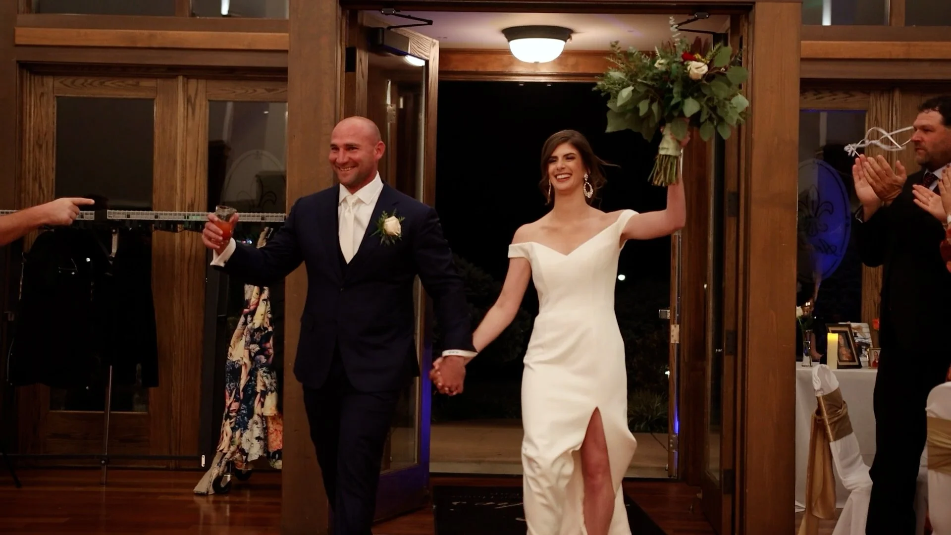 A bride and groom walking hand in hand out of a wedding venue, smiling, with the bride holding a bouquet in her raised hand, while guests applaud in the background.