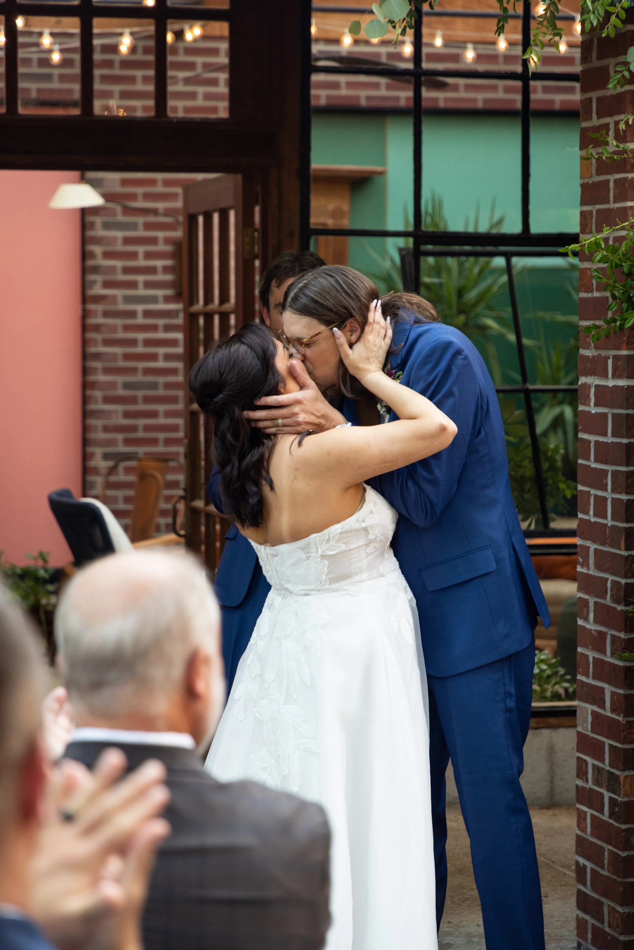 A couple sharing a kiss at a wedding ceremony inside a decorated venue with brick walls, string lights, and large windows, with guests watching.