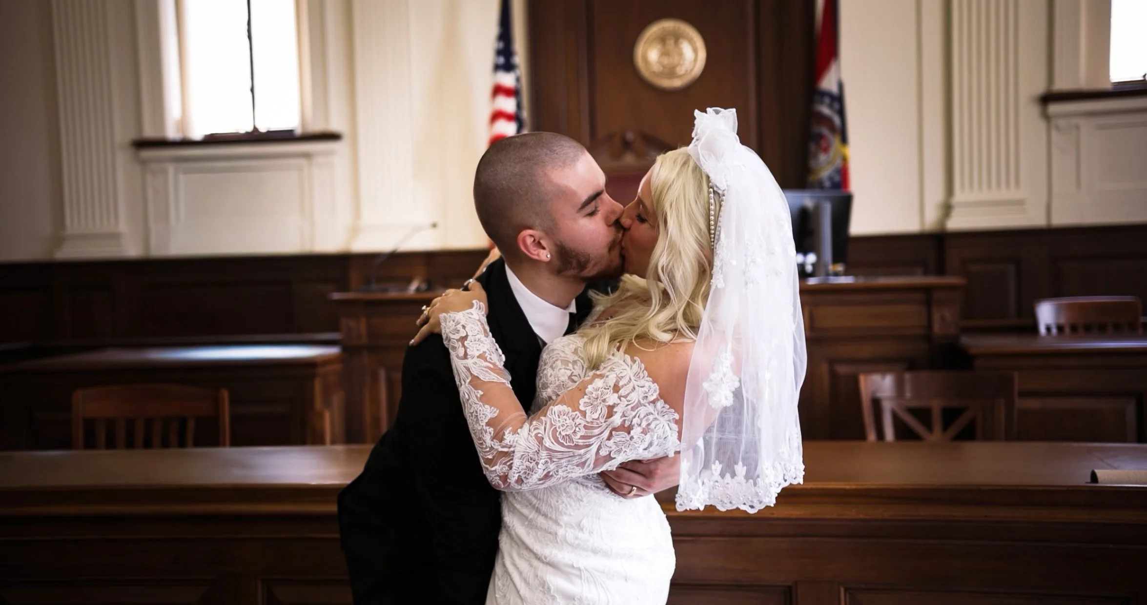 A bride and groom sharing a kiss in a courtroom, with wood-paneled walls, American flag, and judge's bench in the background.