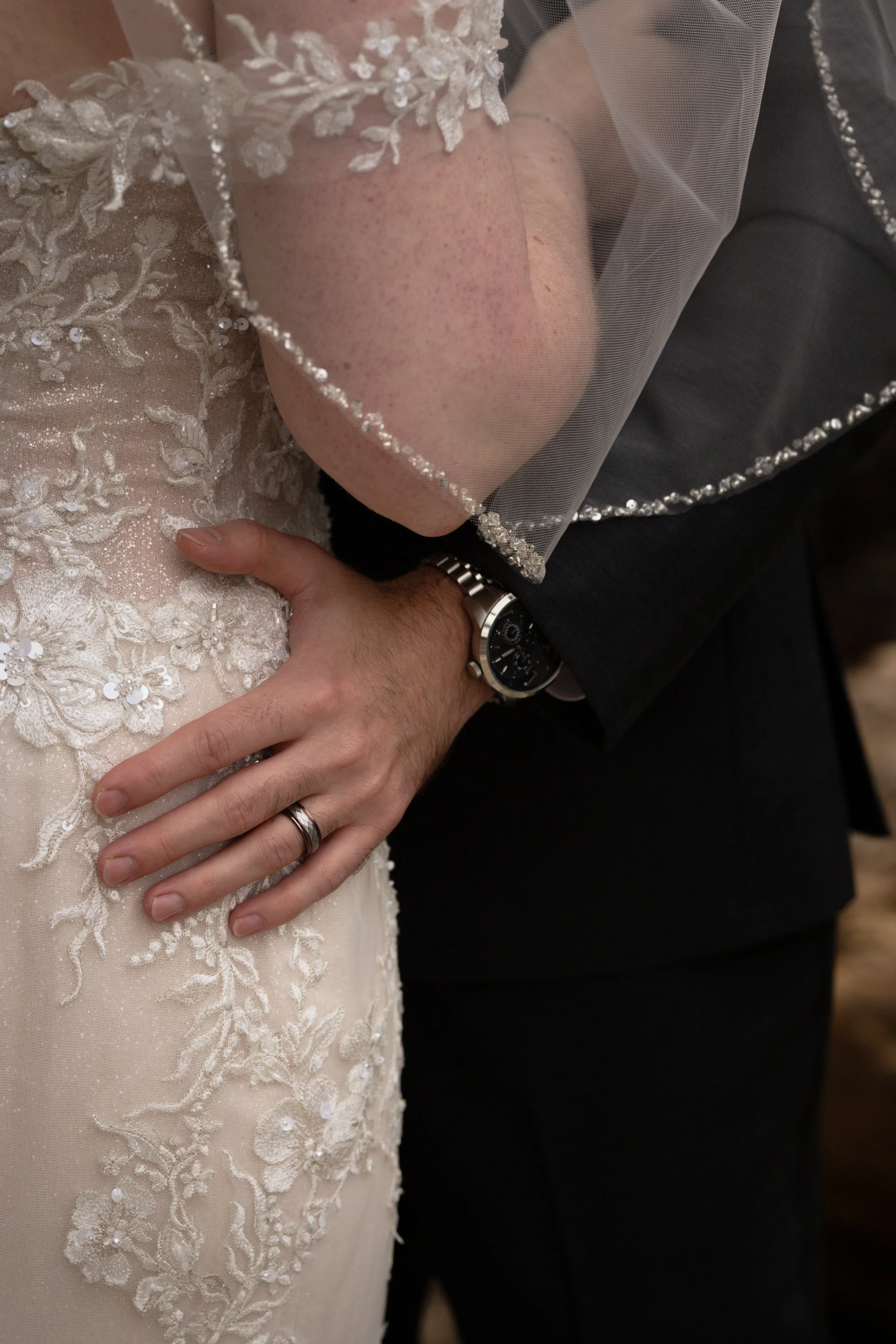 Close-up of a bride and groom embracing at their wedding, focusing on the bride's lace wedding dress, the groom's hand with a wedding band, and the bride's veil with bead detailing.
