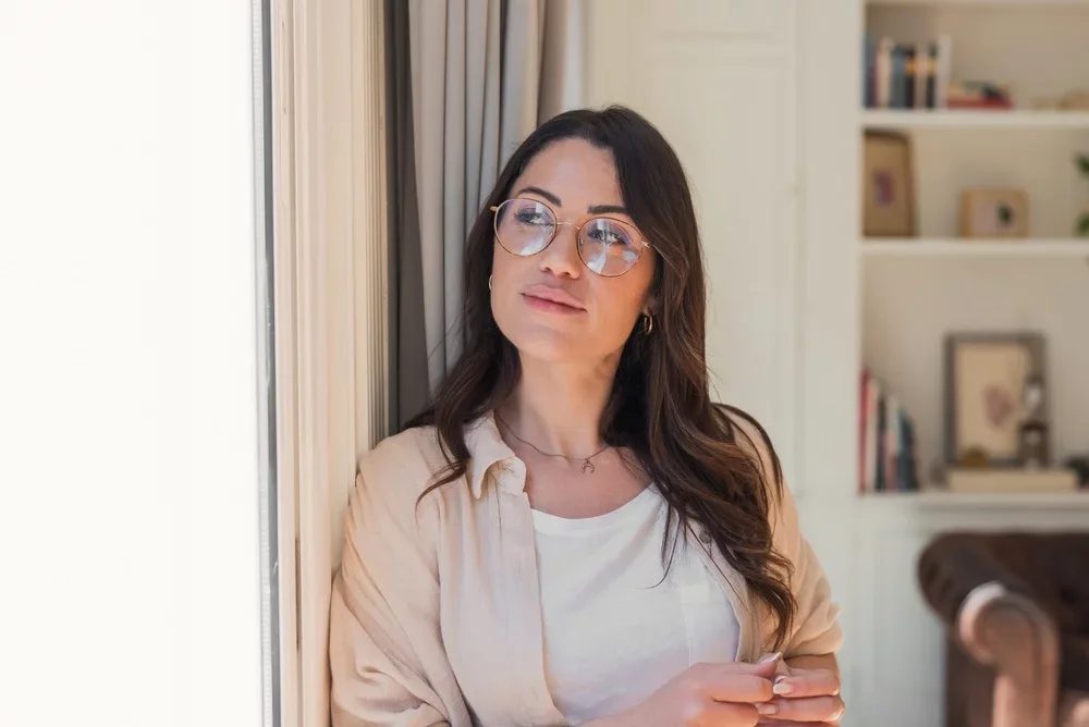 A thoughtful woman wearing glasses gazes out a sunlit window in a cozy home interior.