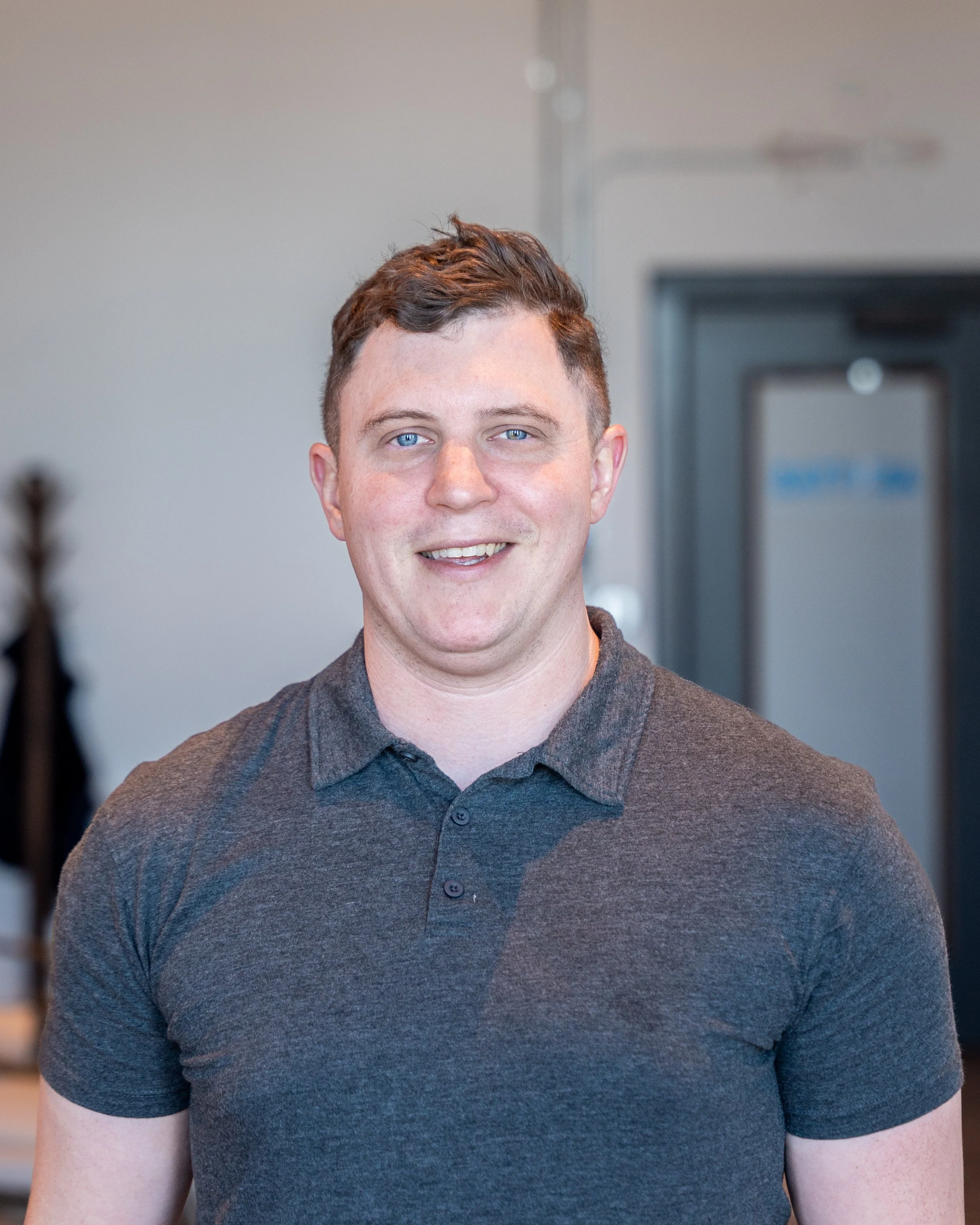 A person wearing a gray polo shirt standing indoors with a blurred background.