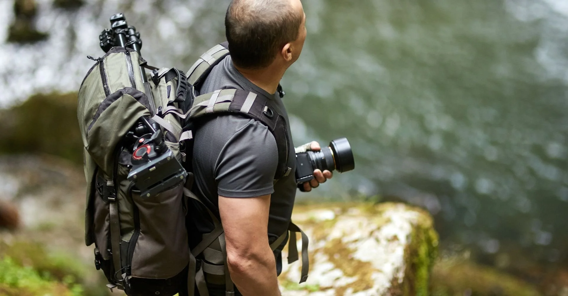 man standing beside a river, camera in hand wearing a backpack with a tripod slung through the top