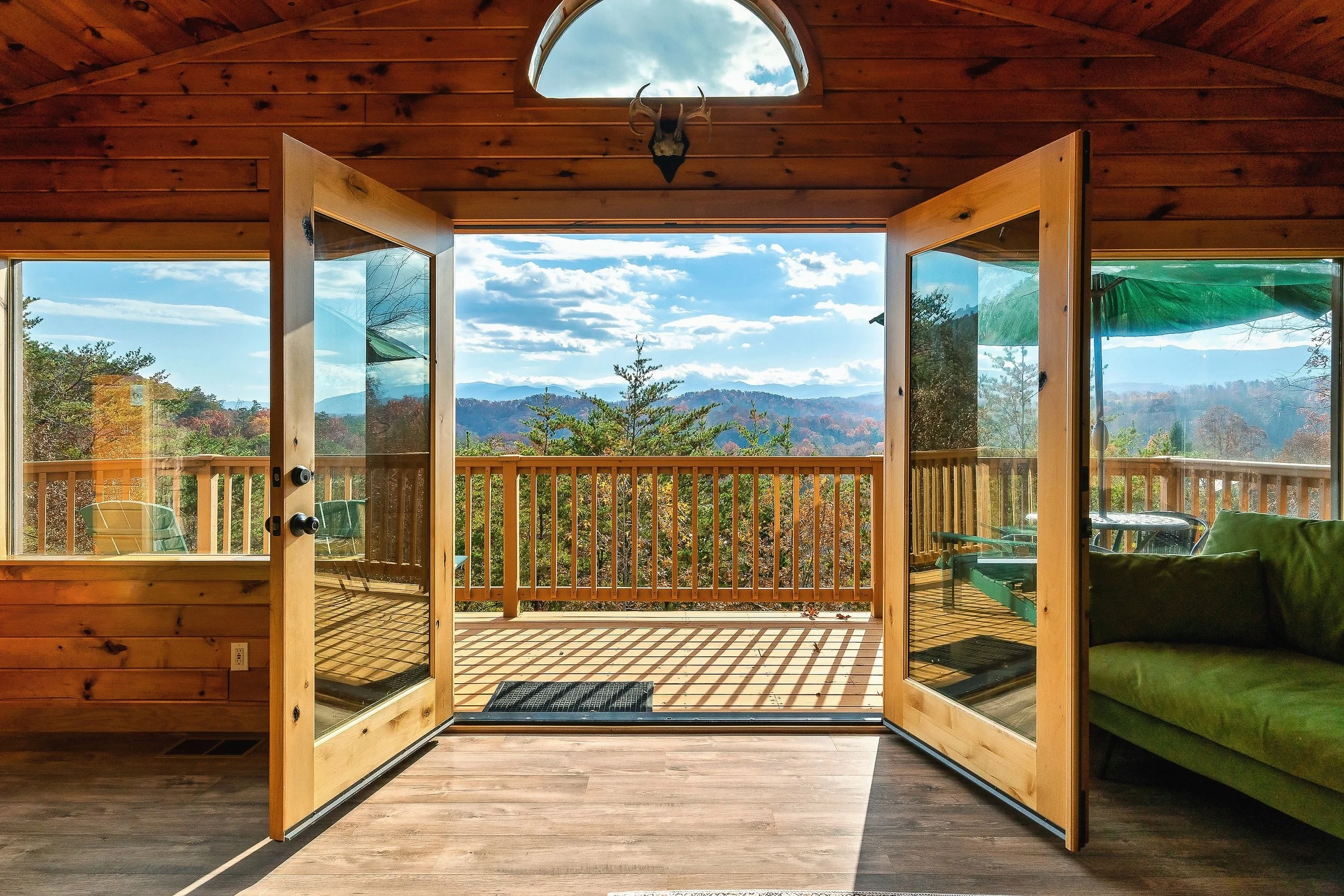Interior view of a wooden cabin with open double glass doors leading to a balcony, showcasing a mountain landscape with clouds, trees, and blue sky.