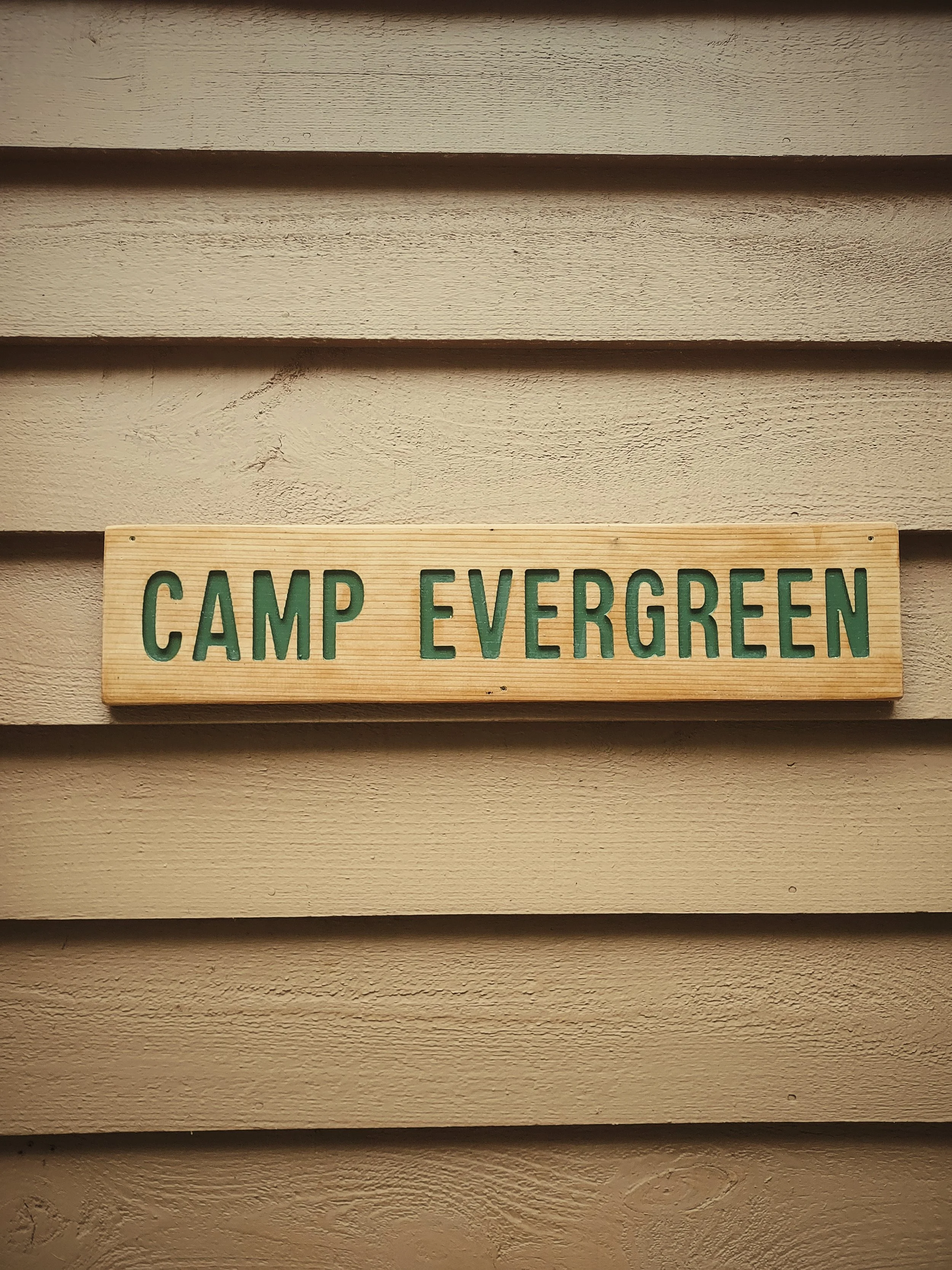 Wooden sign with green text reading 'CAMP EVERGREEN' mounted on a beige horizontal wooden siding wall.