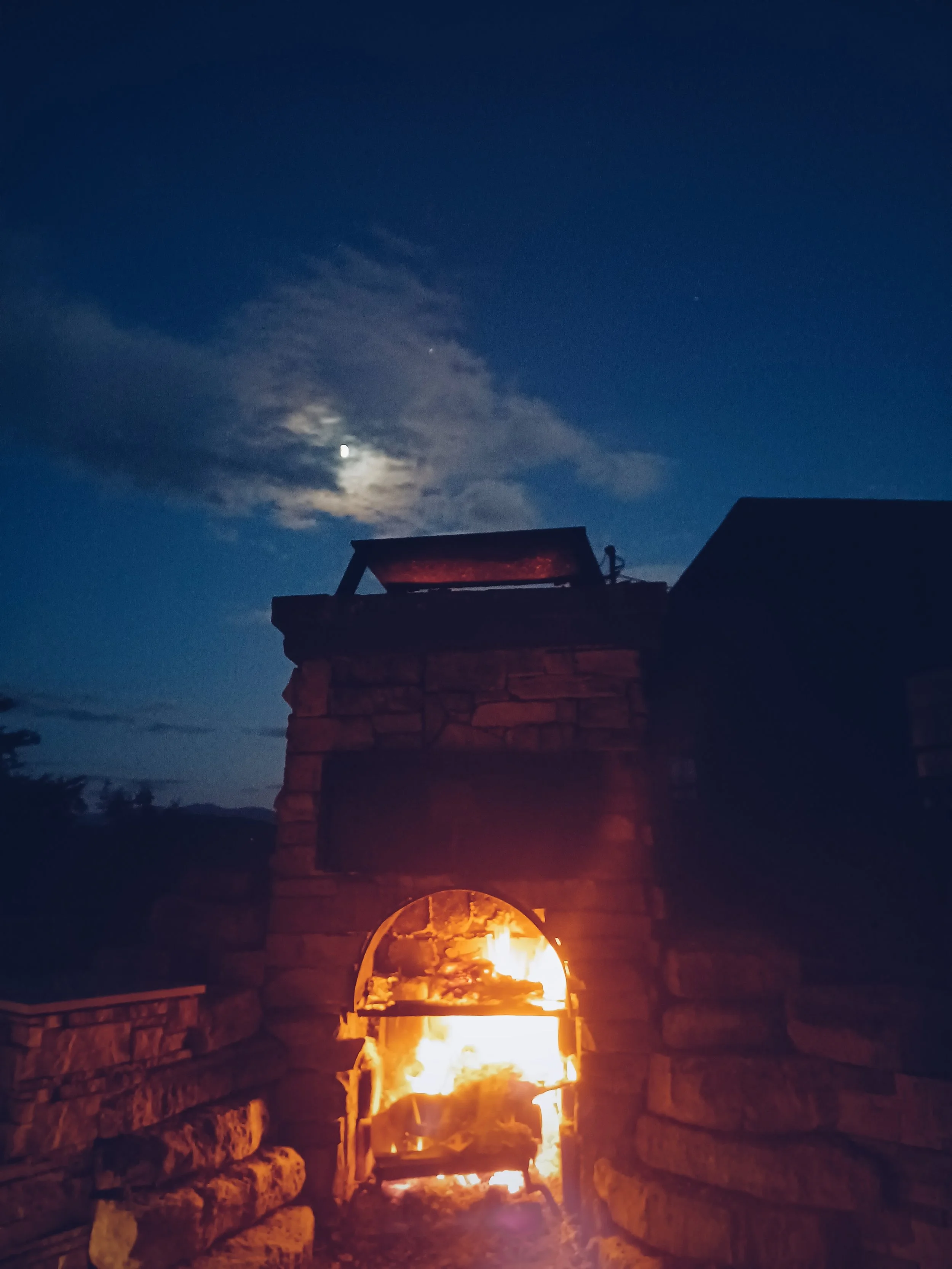 A stone fireplace with a fire burning inside, set against a dark evening sky with scattered clouds and visible stars.