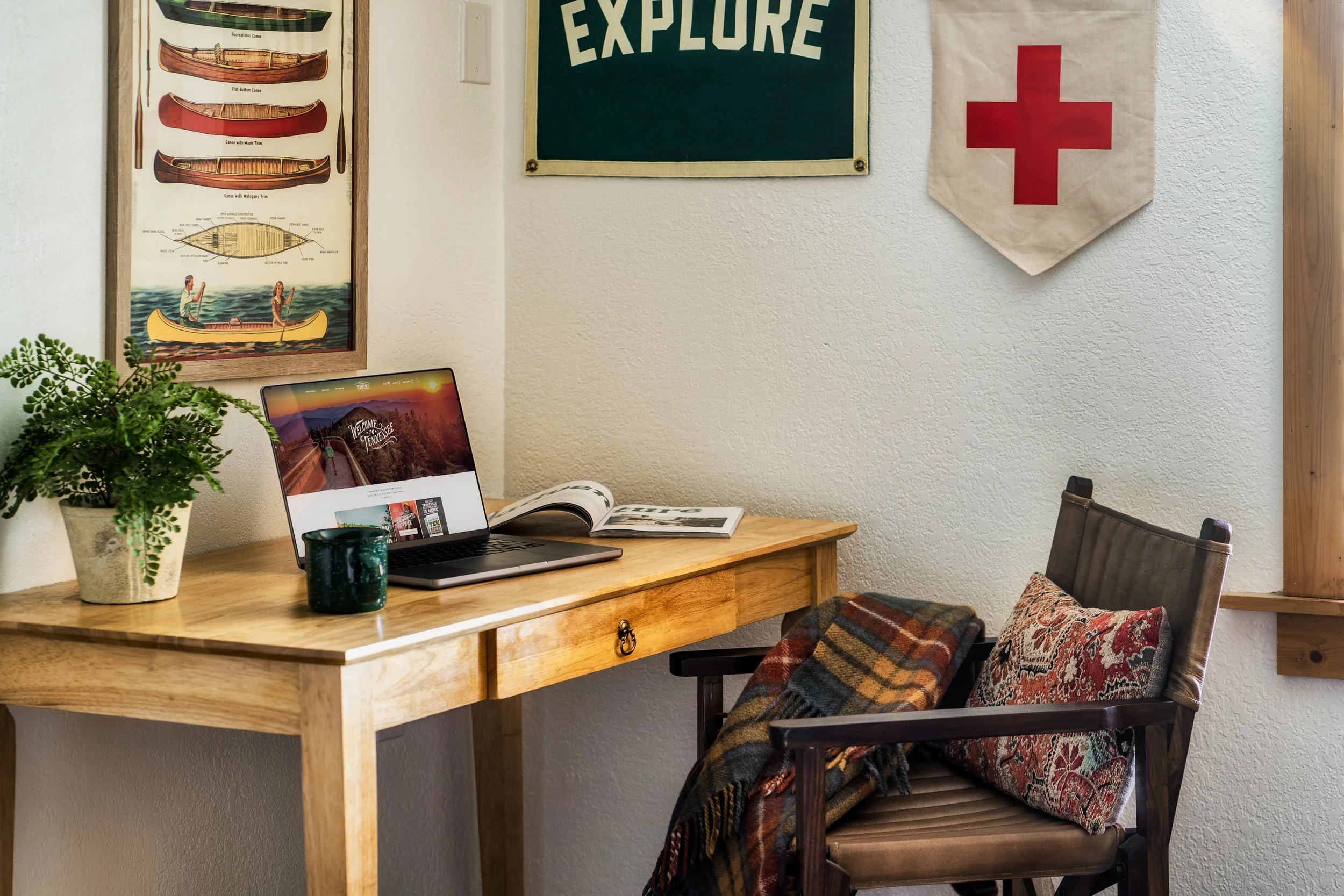 A workspace with a wooden desk and a chair, decorated with a potted plant, a computer displaying a Tennessee website, an open magazine, and a green mug. On the wall, there are framed canoe diagrams, a green sign with the word 'EXPLORE,' and a Red Cro