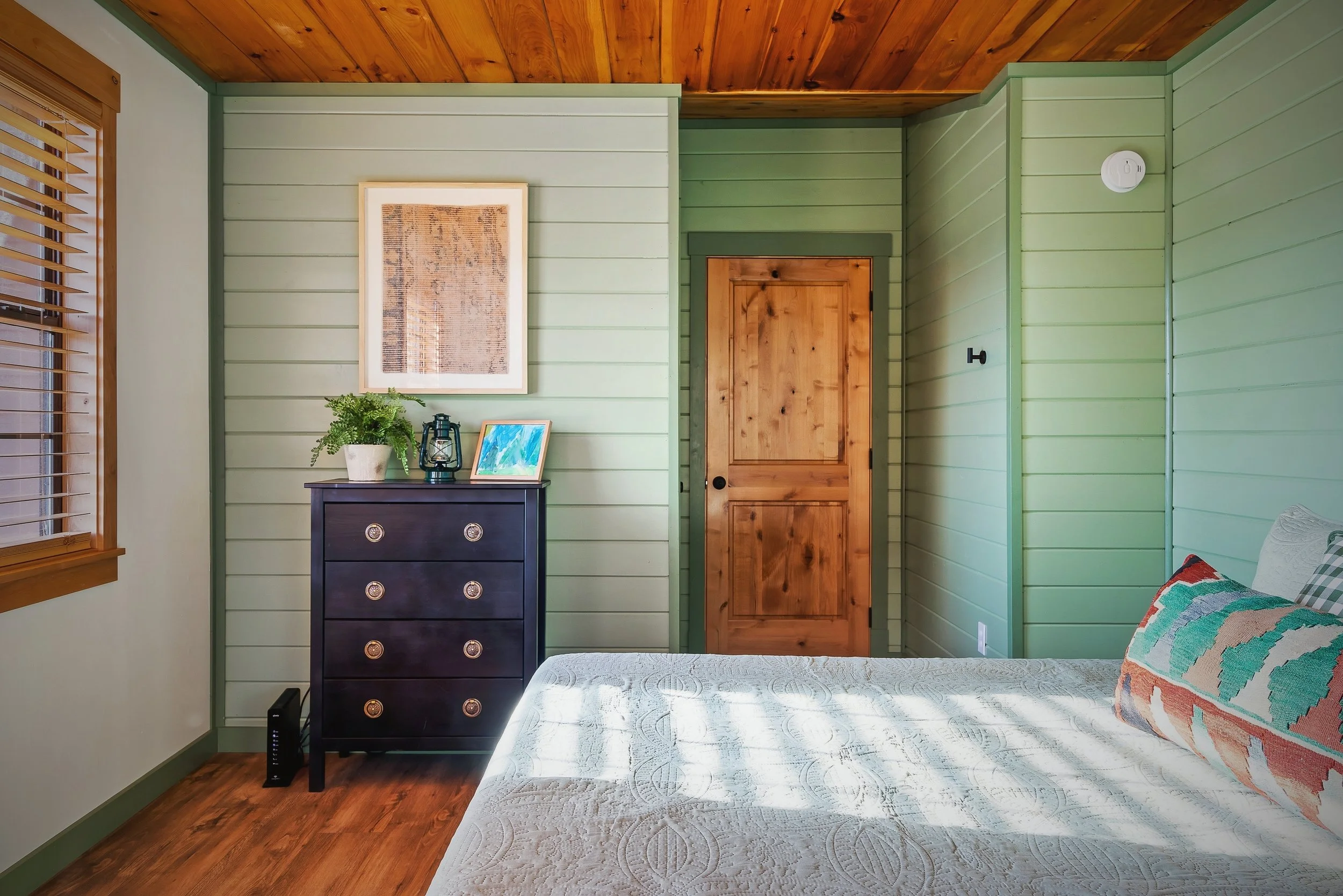 A bedroom with green painted wood panel walls, a wooden ceiling, a window with wooden blinds, a black dresser with decorative knobs, a picture frame, a plant, a lantern, a bed with a white quilted bedspread and colorful pillows, and a wooden door.