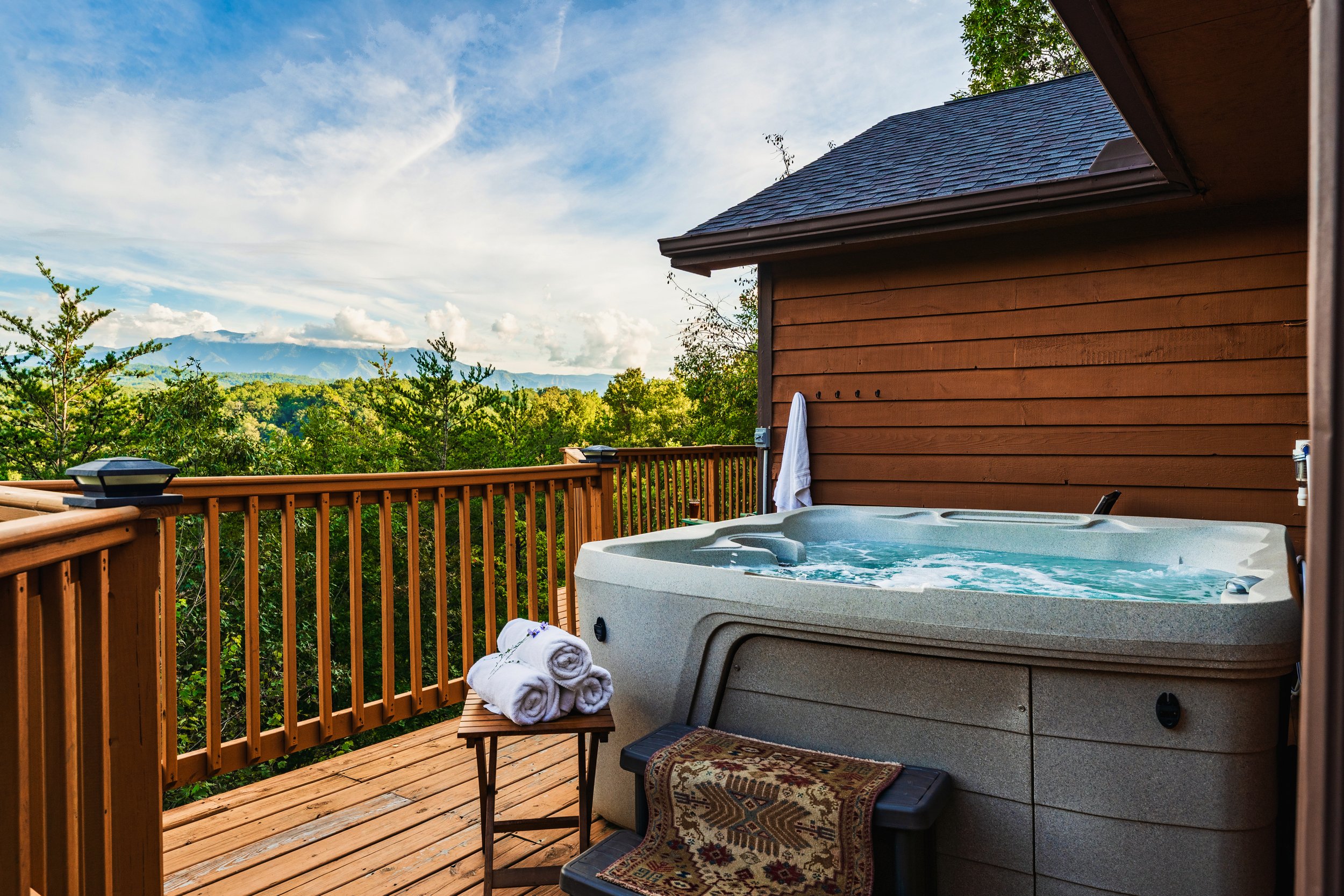 A hot tub on a wooden deck with rolled towels on a small table, overlooking a lush green forest and mountains in the distance, under a partly cloudy sky.