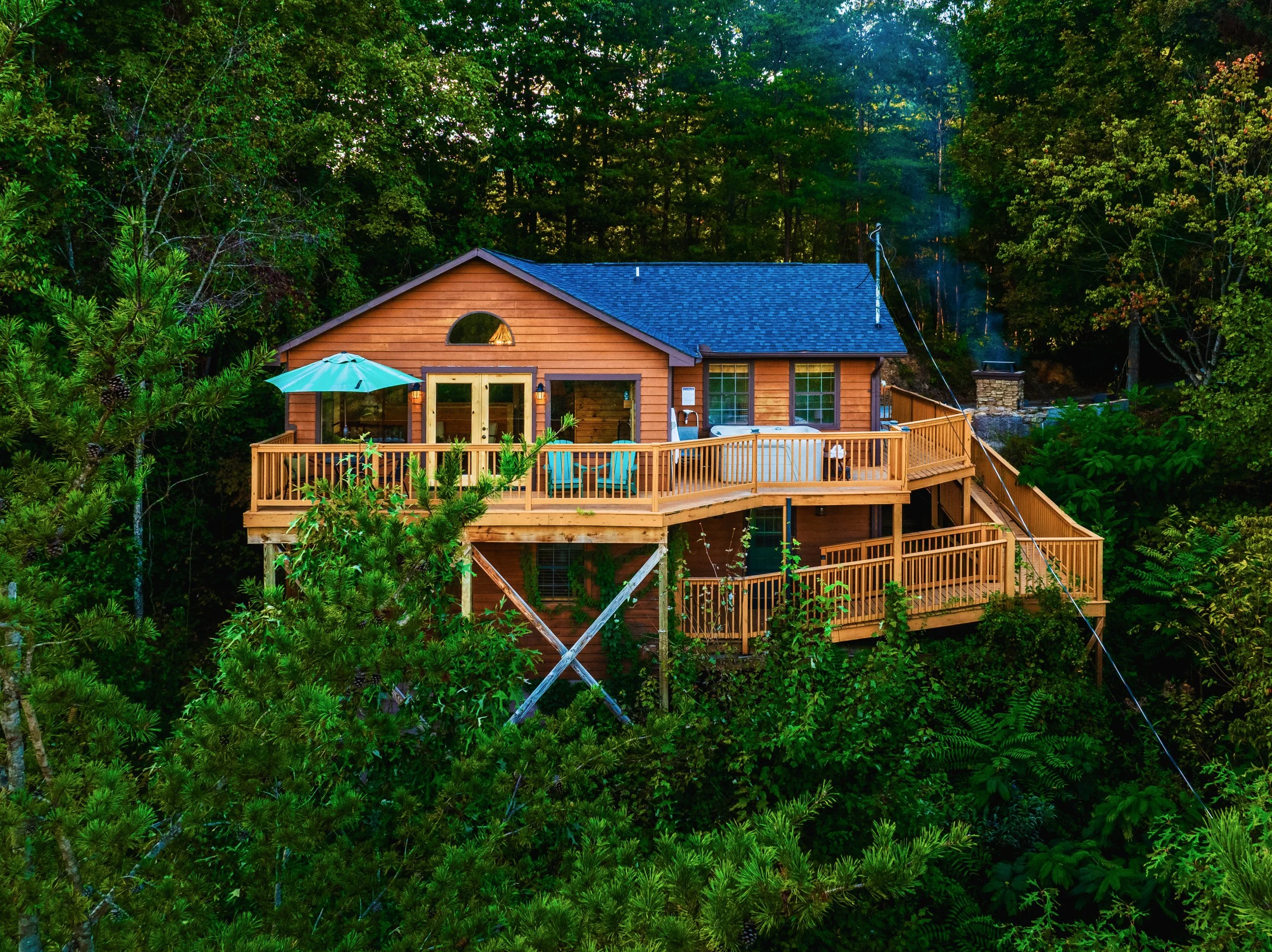 A wooden house with a deck and an umbrella set outdoors amidst a dense forest with lush green trees.