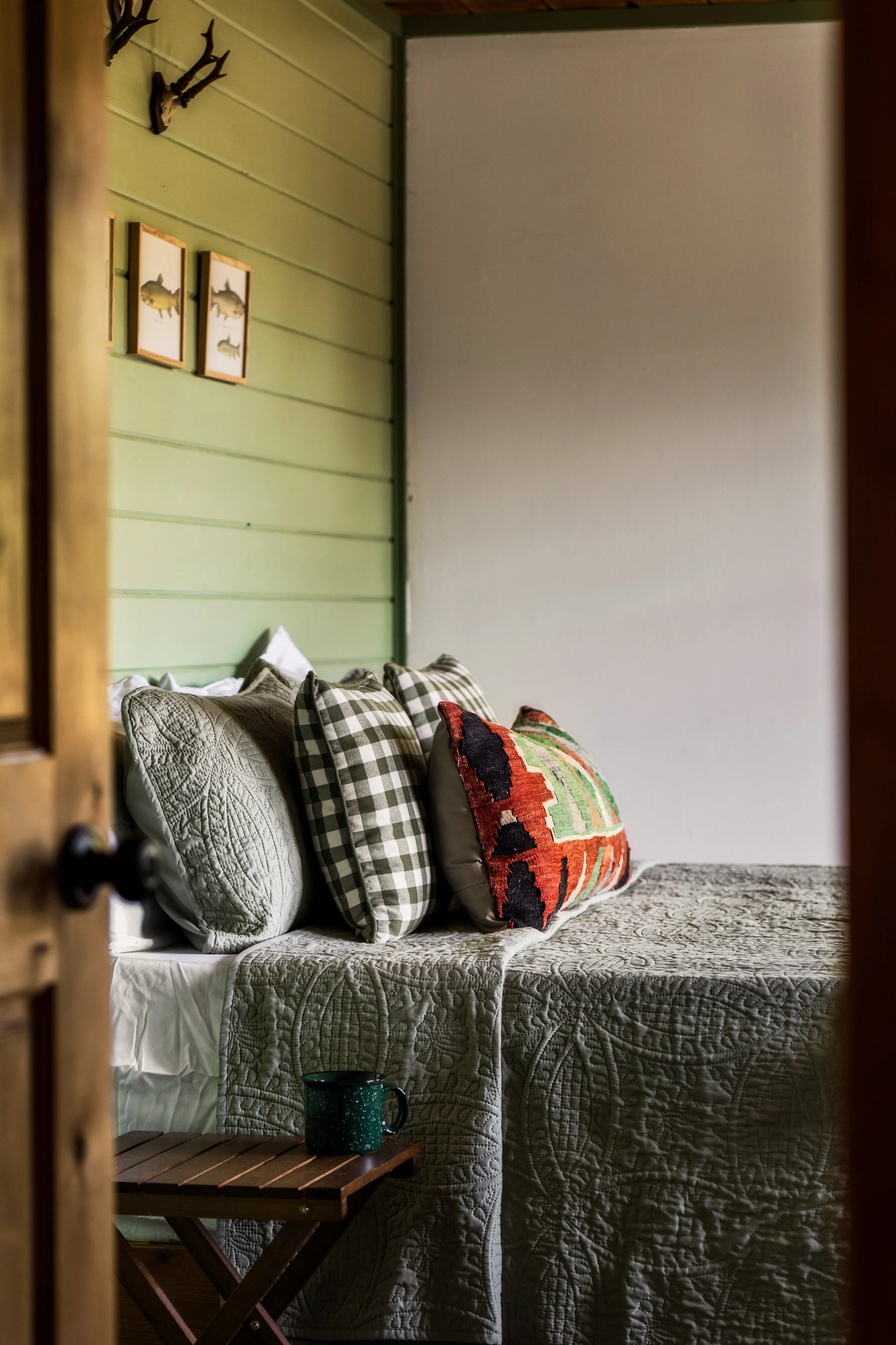 A cozy bed with a quilt and pillows, including checkered and patterned designs, against a green paneled wall with framed fish artwork and antler decor, with a small wooden table holding a green mug.