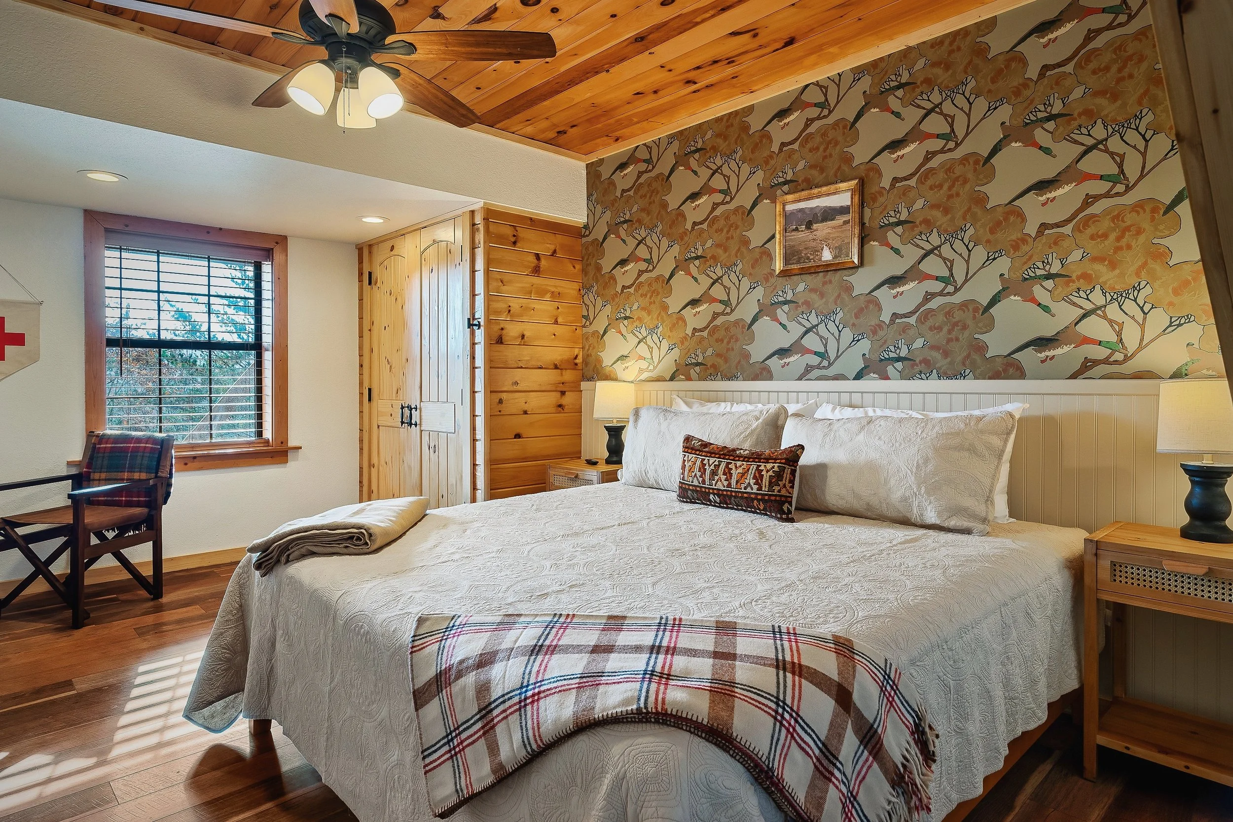 A cozy bedroom with a wooden ceiling, wallpaper with a bird and tree pattern, a bed with white quilt, plaid blanket, and pillows, pine furniture, a window with blinds, a ceiling fan with lights, and a framed landscape picture on the wall.