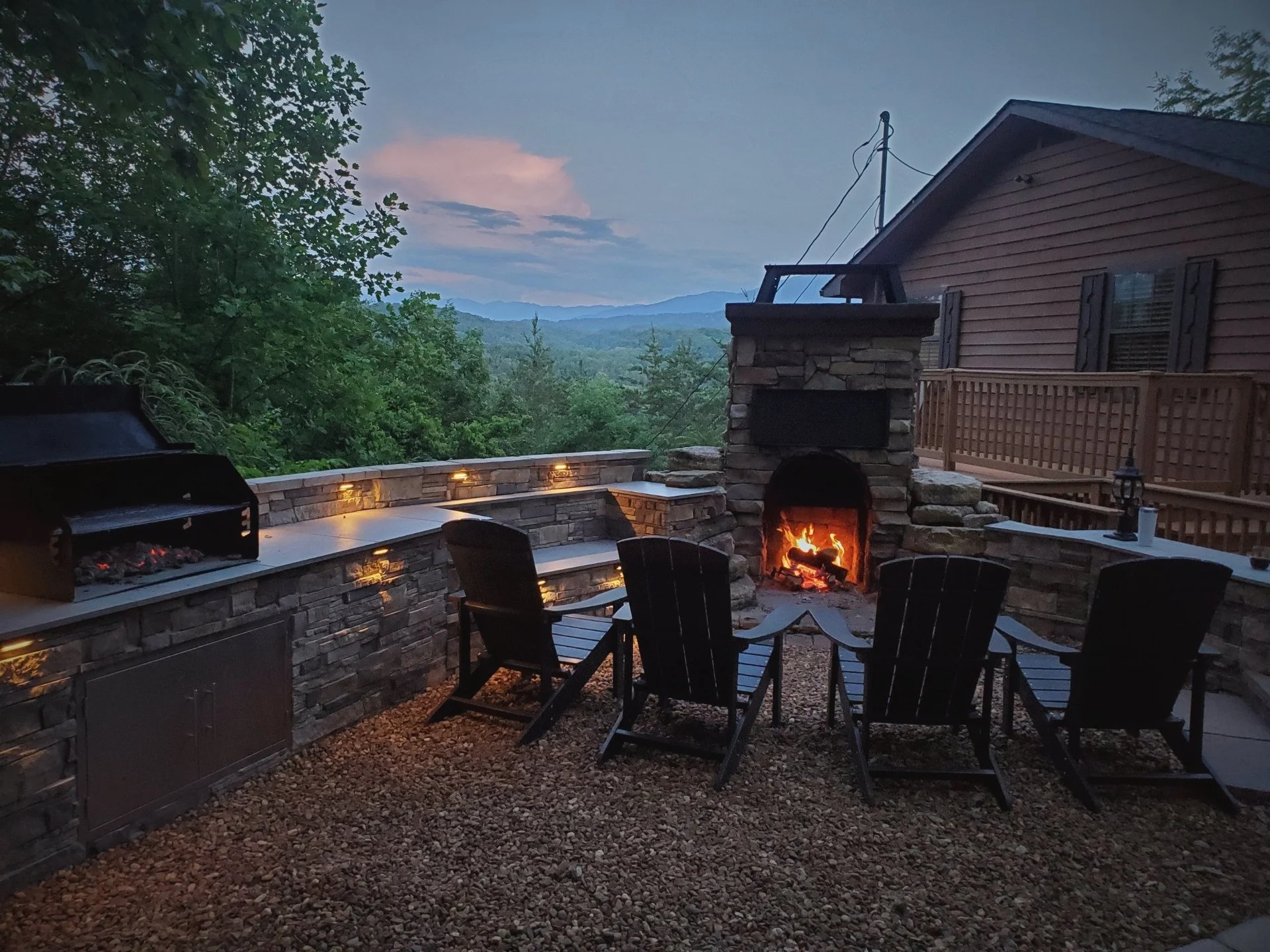Outdoor backyard deck featuring a stone fireplace with a fire burning, surrounded by chairs, with a view of mountains in the distance during dusk.