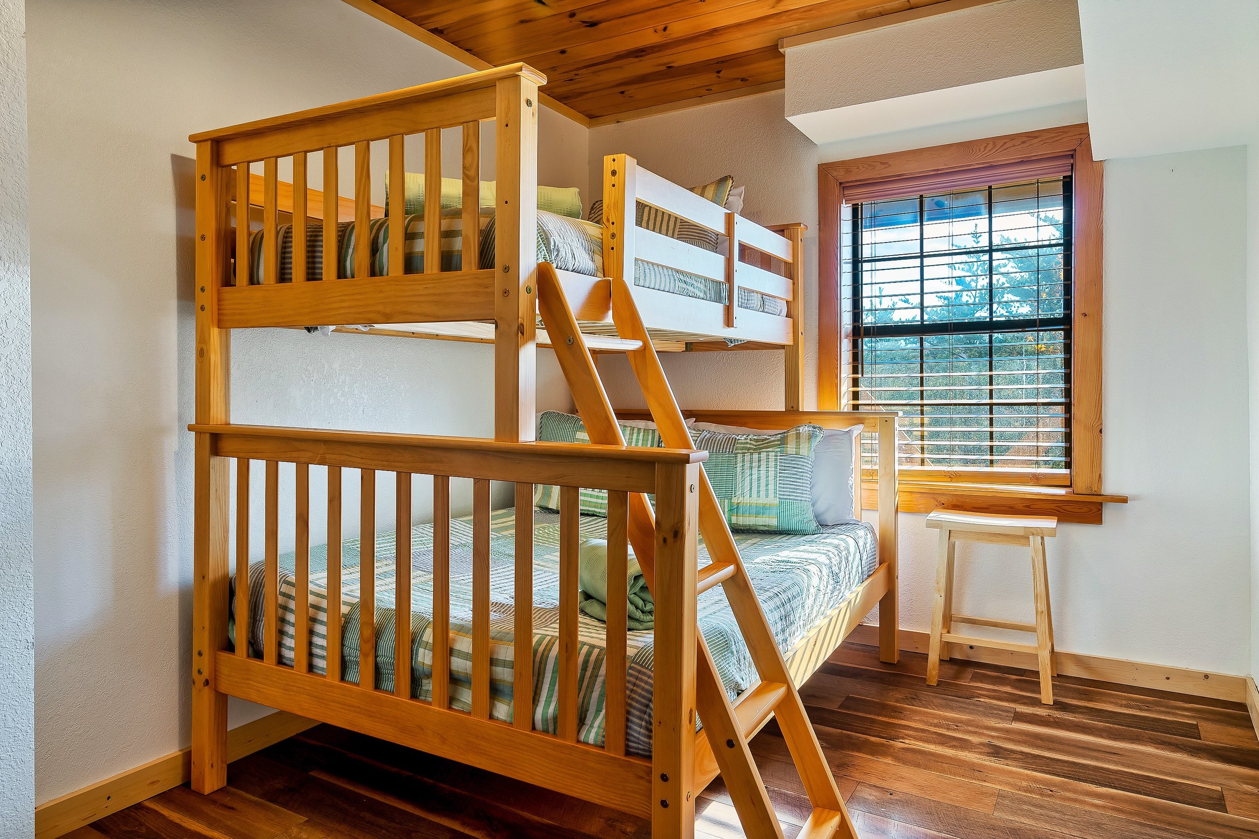 Wooden bunk beds with striped bedding and pillows in a cozy room with wooden floor, walls, and ceiling, and a large window with blinds showing trees outside.