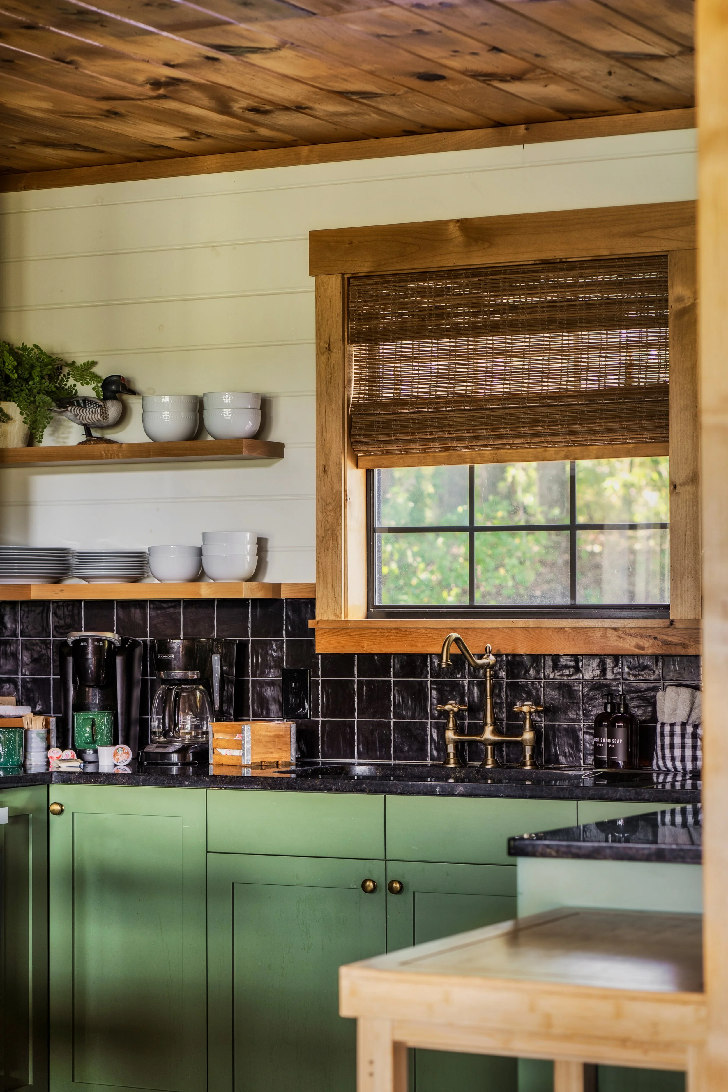 A cozy kitchen with black tile backsplash, green cabinets, and open shelves holding white bowls and a decorative duck figurine, with a window covered by a bamboo shade.