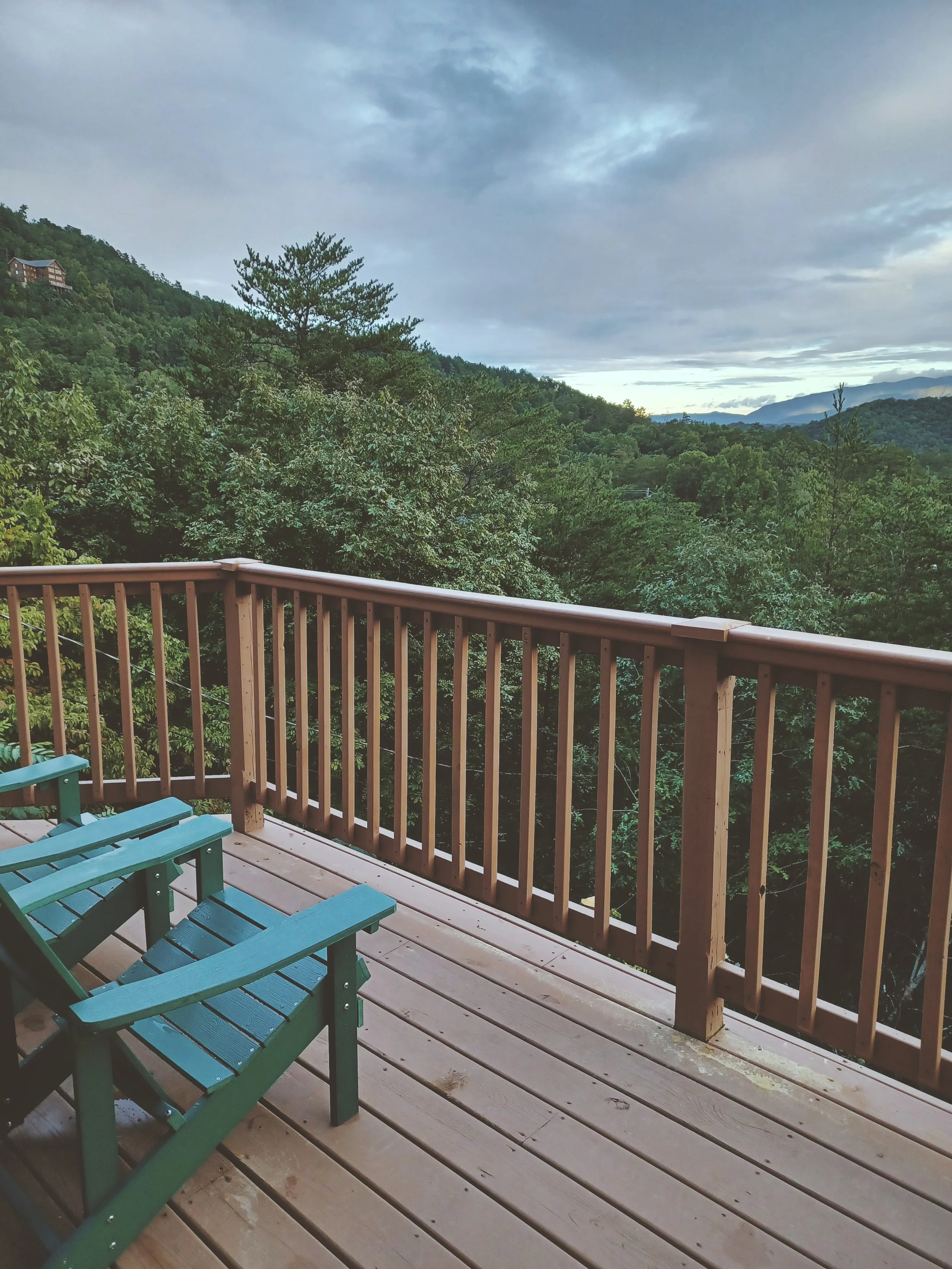 A wooden deck with a railing and green Adirondack chairs, overlooking a lush, forested mountain landscape under a cloudy sky.