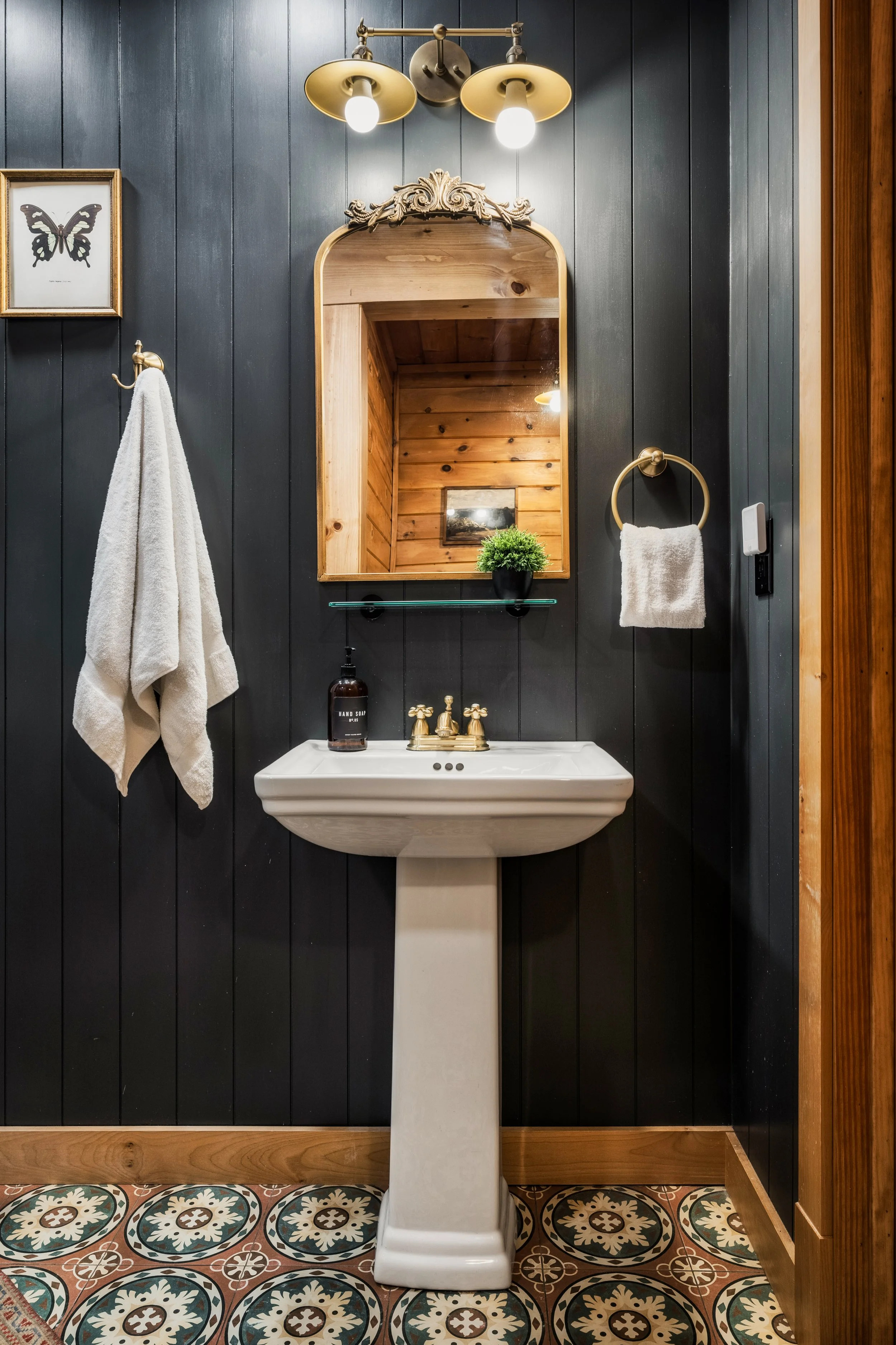 A bathroom with a black wood-paneled wall, a white sink with vintage brass fixtures, a large mirror with ornate wood trim, a small potted plant on a glass shelf, a white towel hanging on a gold ring, a framed butterfly illustration, and patterned til