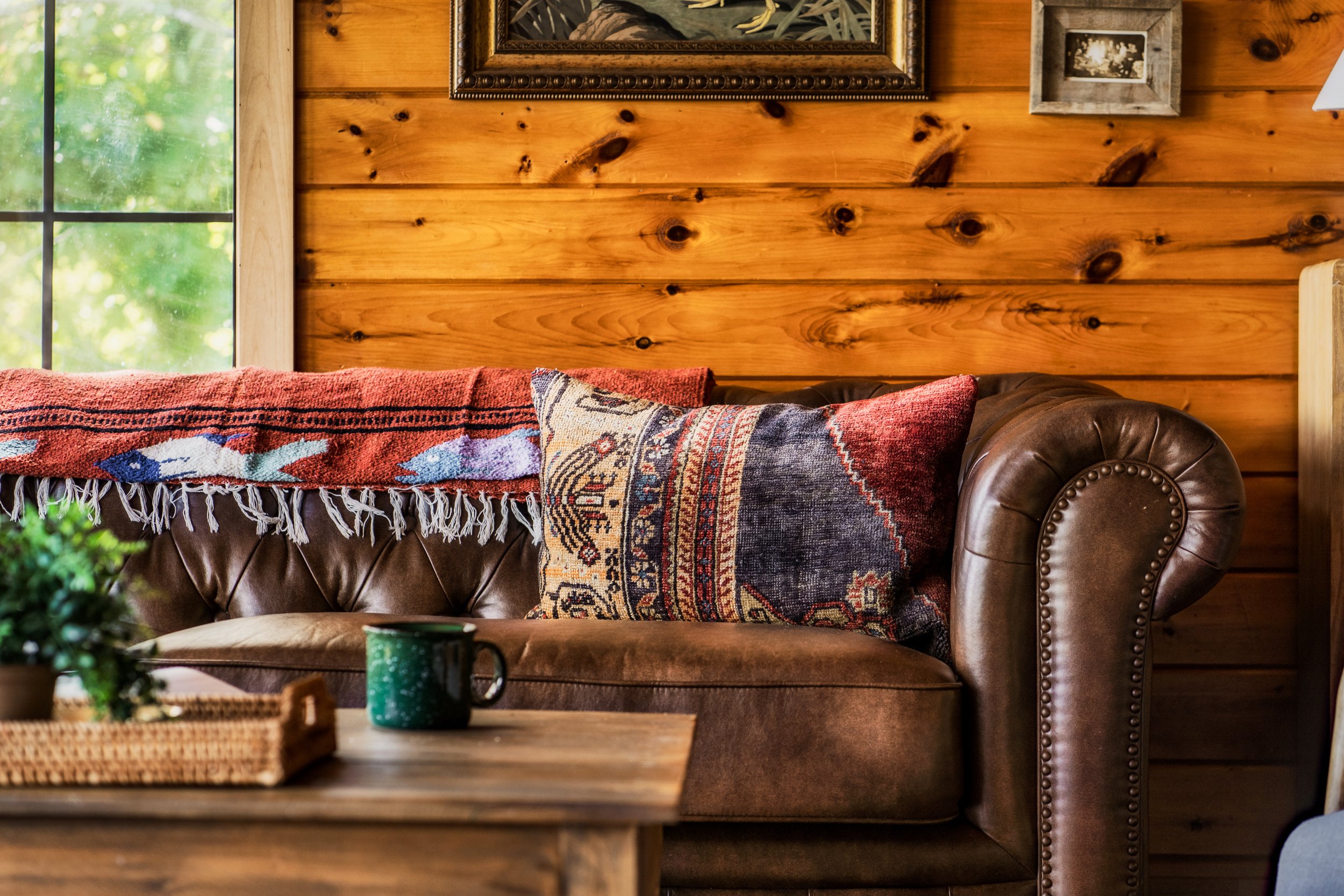 A cozy cabin living room with a brown leather sofa, colorful patterned pillows, a wooden coffee table with a green cup and a basket, wood-paneled walls, and framed artwork. A window showing greenery outside is on the left.
