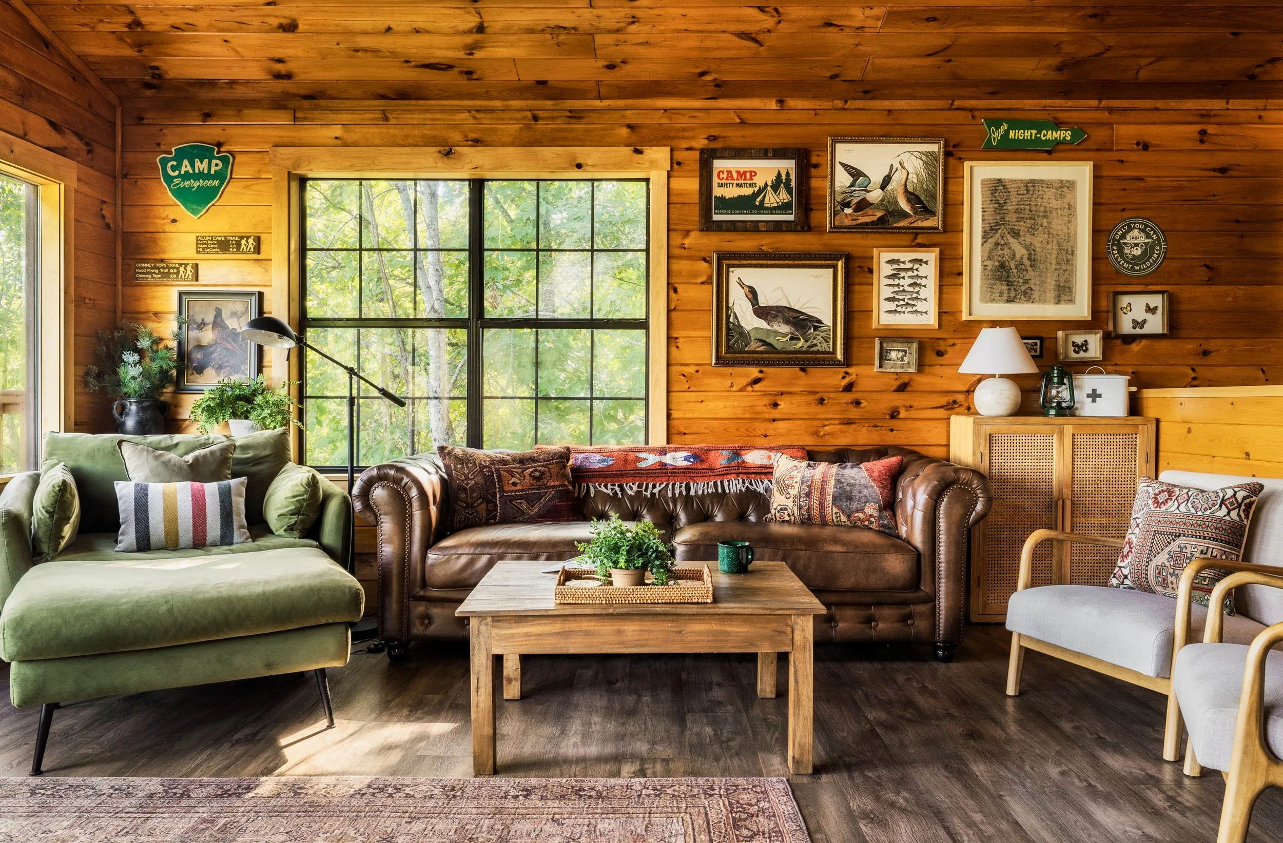 Living room with wooden walls and large window overlooking trees, decorated with nature-themed artwork, a green armchair, a brown leather sofa, side table with plants, lamp, and framed pictures.