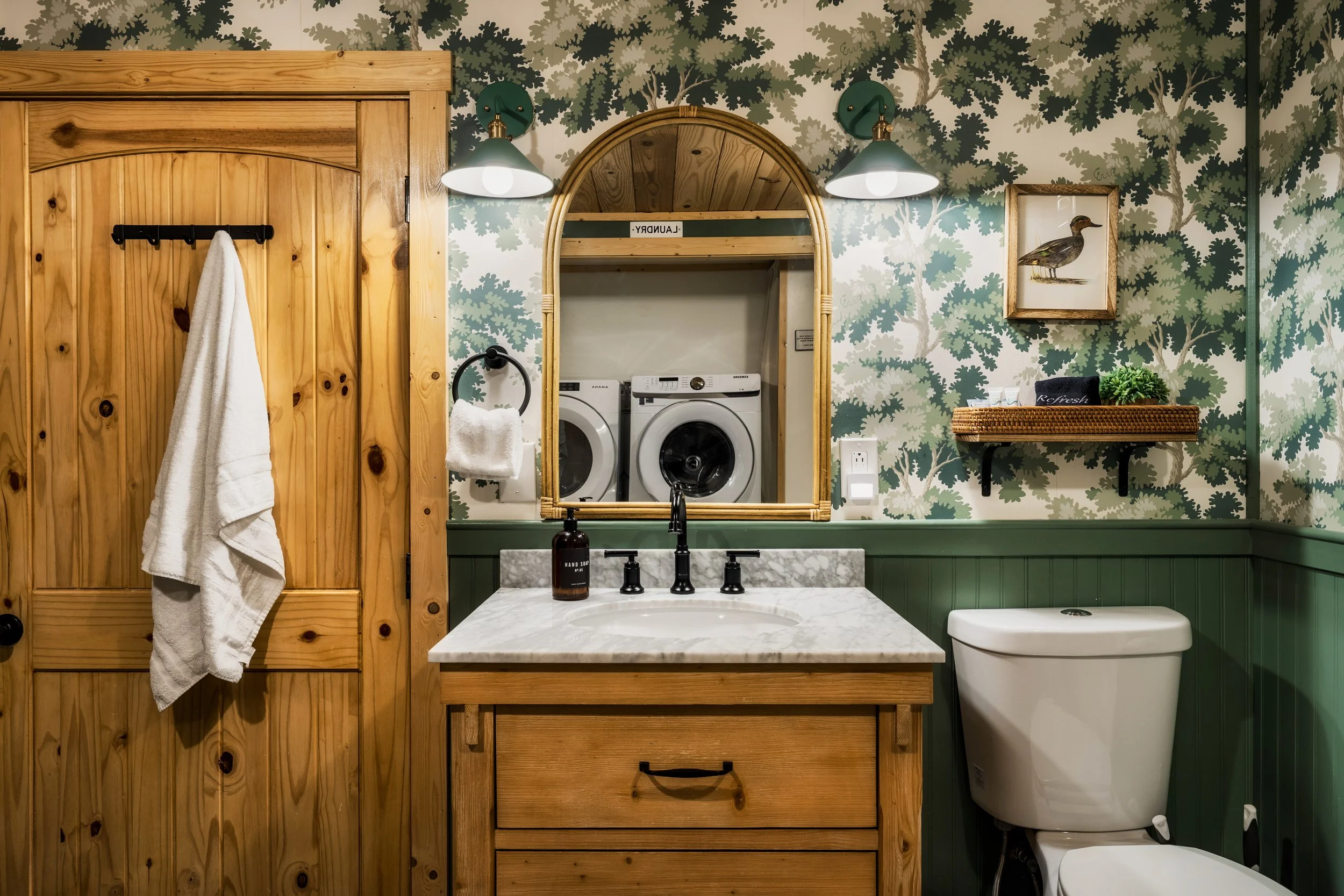 Bathroom with wood and green accents, featuring a vanity with a marble countertop, a framed picture of a duck, and a washing machine visible through an arched mirror opening.