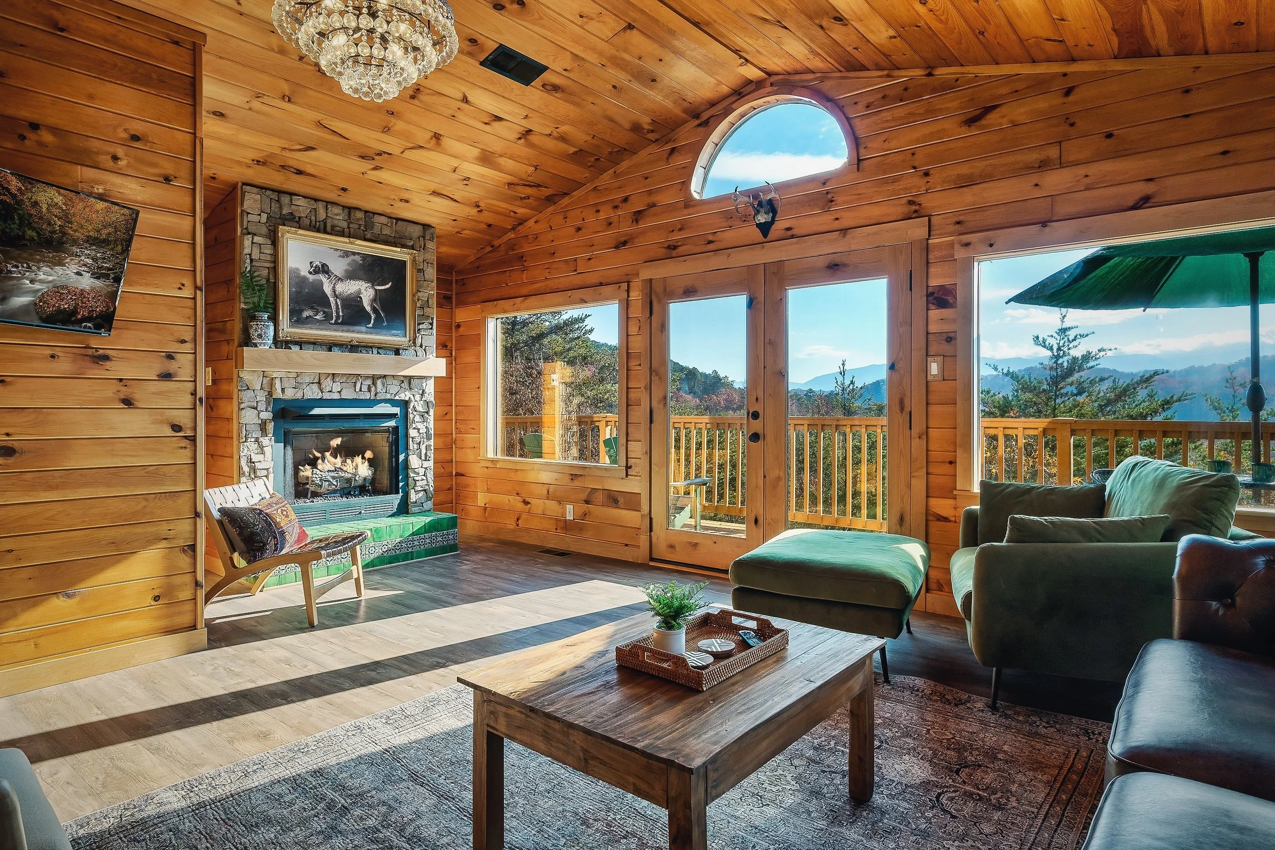 Living room with wooden walls and ceiling, a stone fireplace with a dog painting above, windows showing mountain and forest views, a green umbrella outside, a chandelier, a TV, and patio chairs.